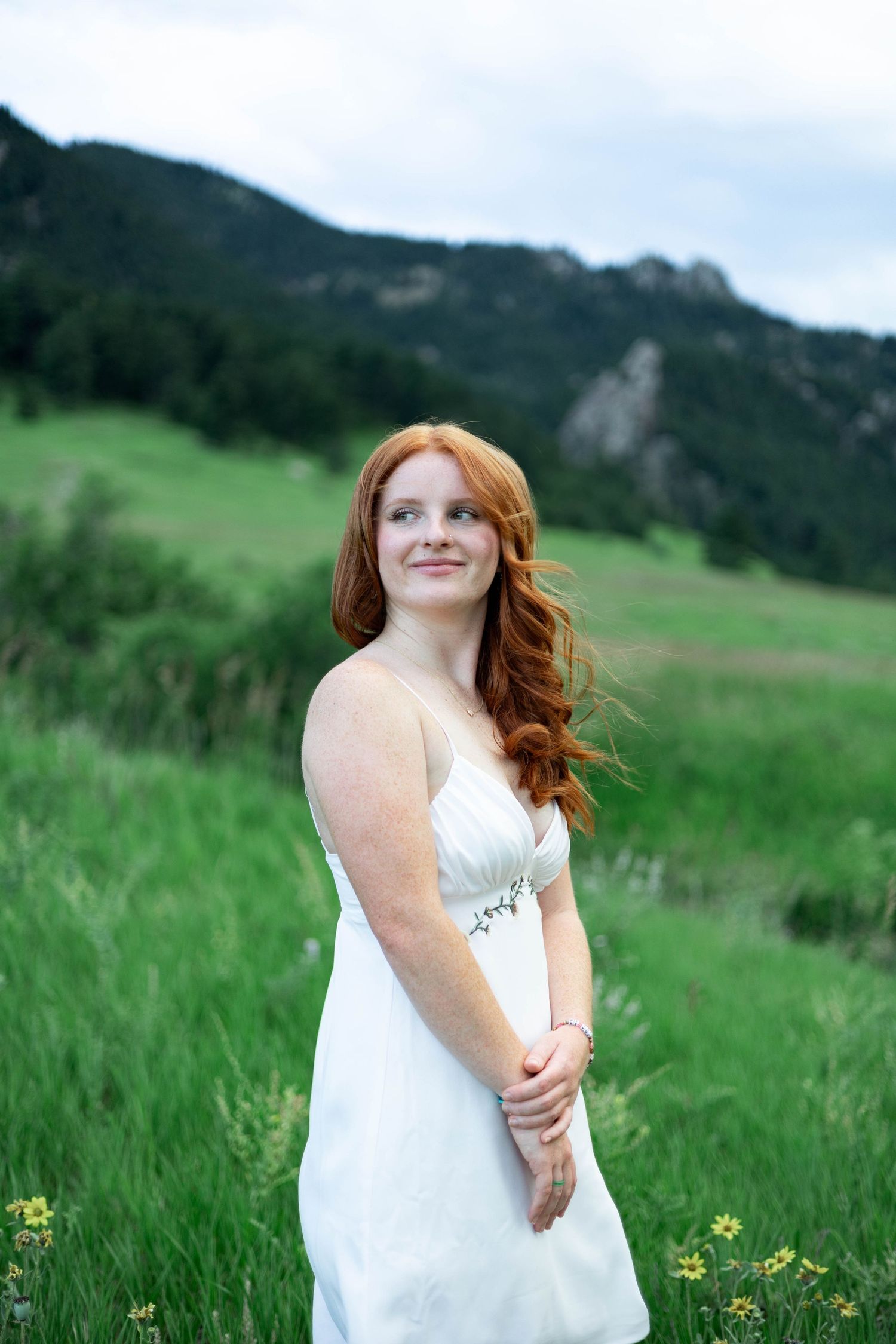 A person in a white dress stands in a green mountain meadow with wildflowers on a summer day.