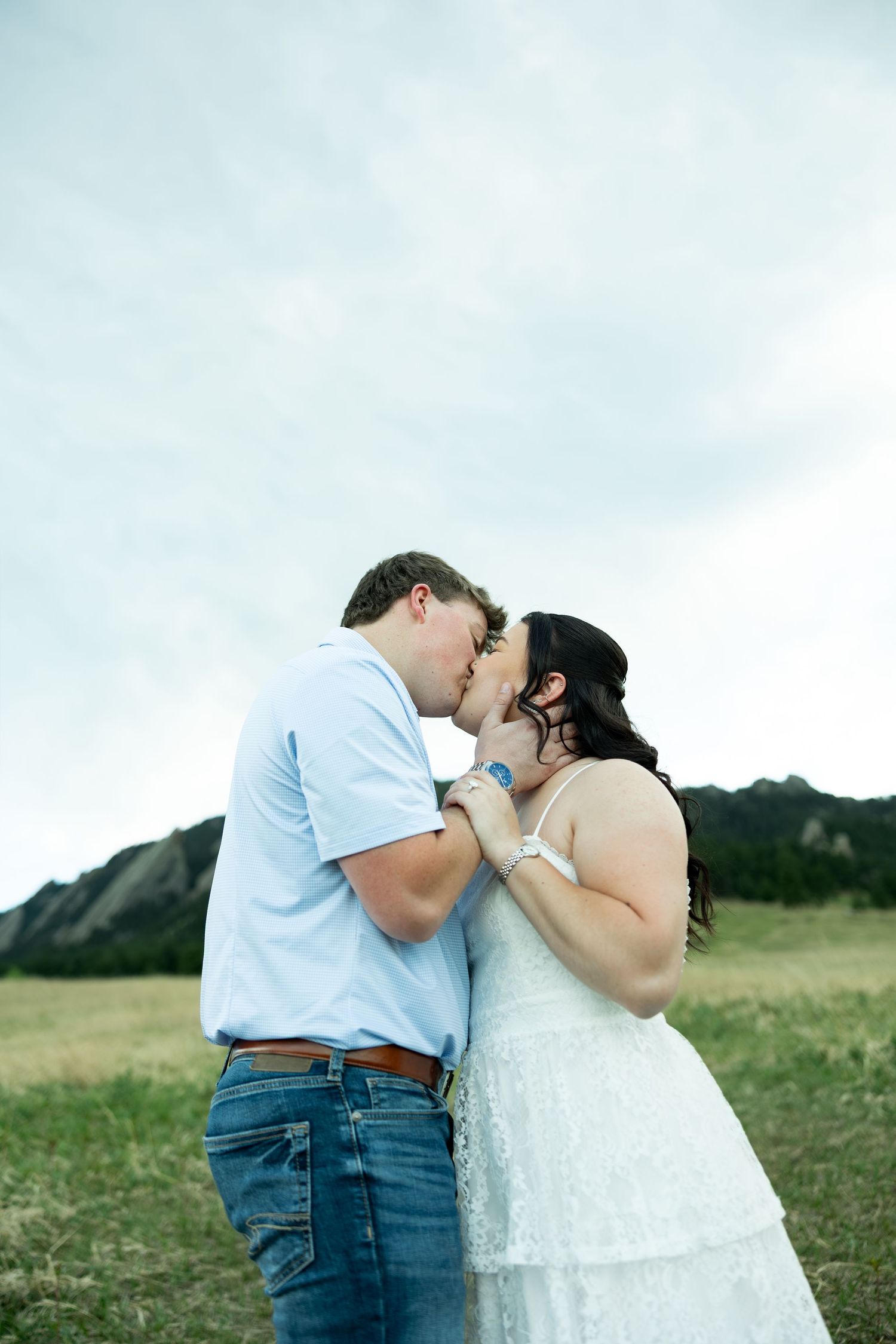 Couple sharing a romantic kiss in a field with rocky mountain flatirons backdrop under a cloudy sky.
