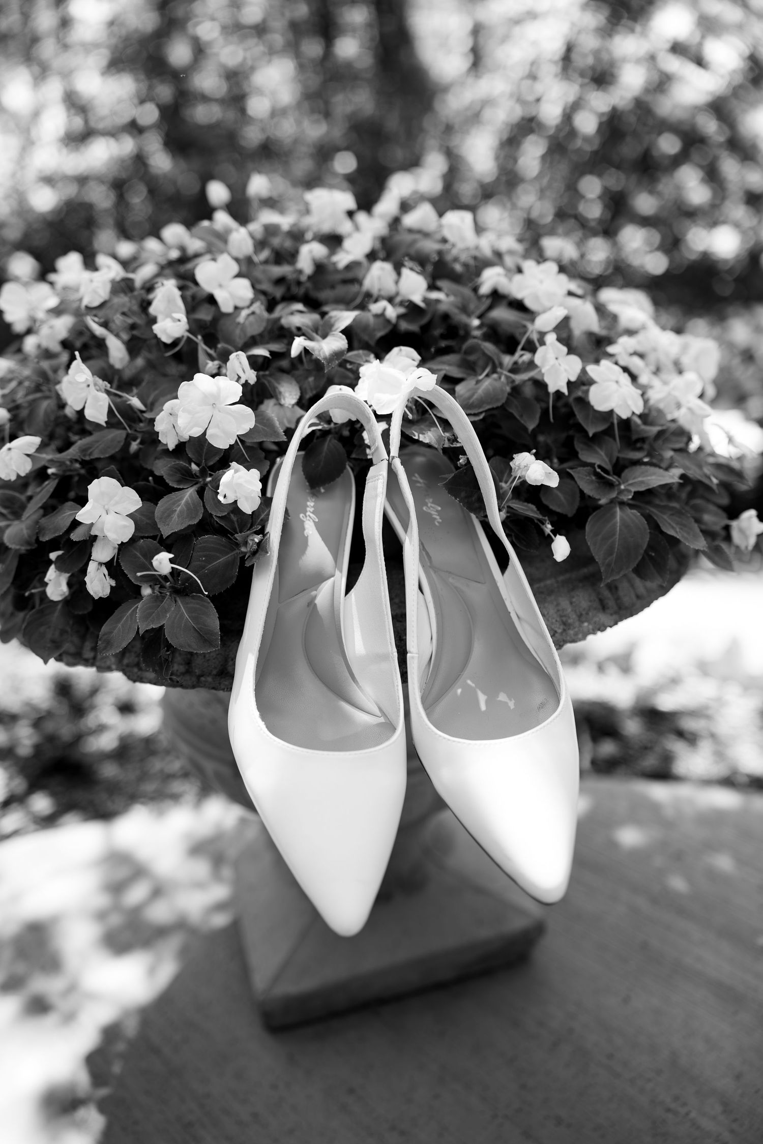 Elegant white bridal heels displayed in front of a floral arrangement, captured in black and white photography.