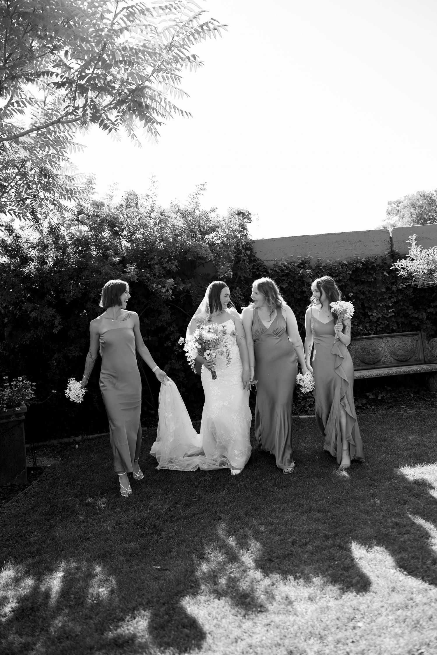 Bride and three bridesmaids in elegant gowns walking outdoors in black and white photo.
