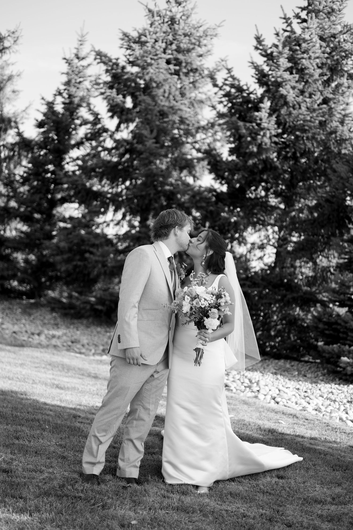 Black and white photo showing backs of people in wedding attire standing by evergreen trees outdoors.
