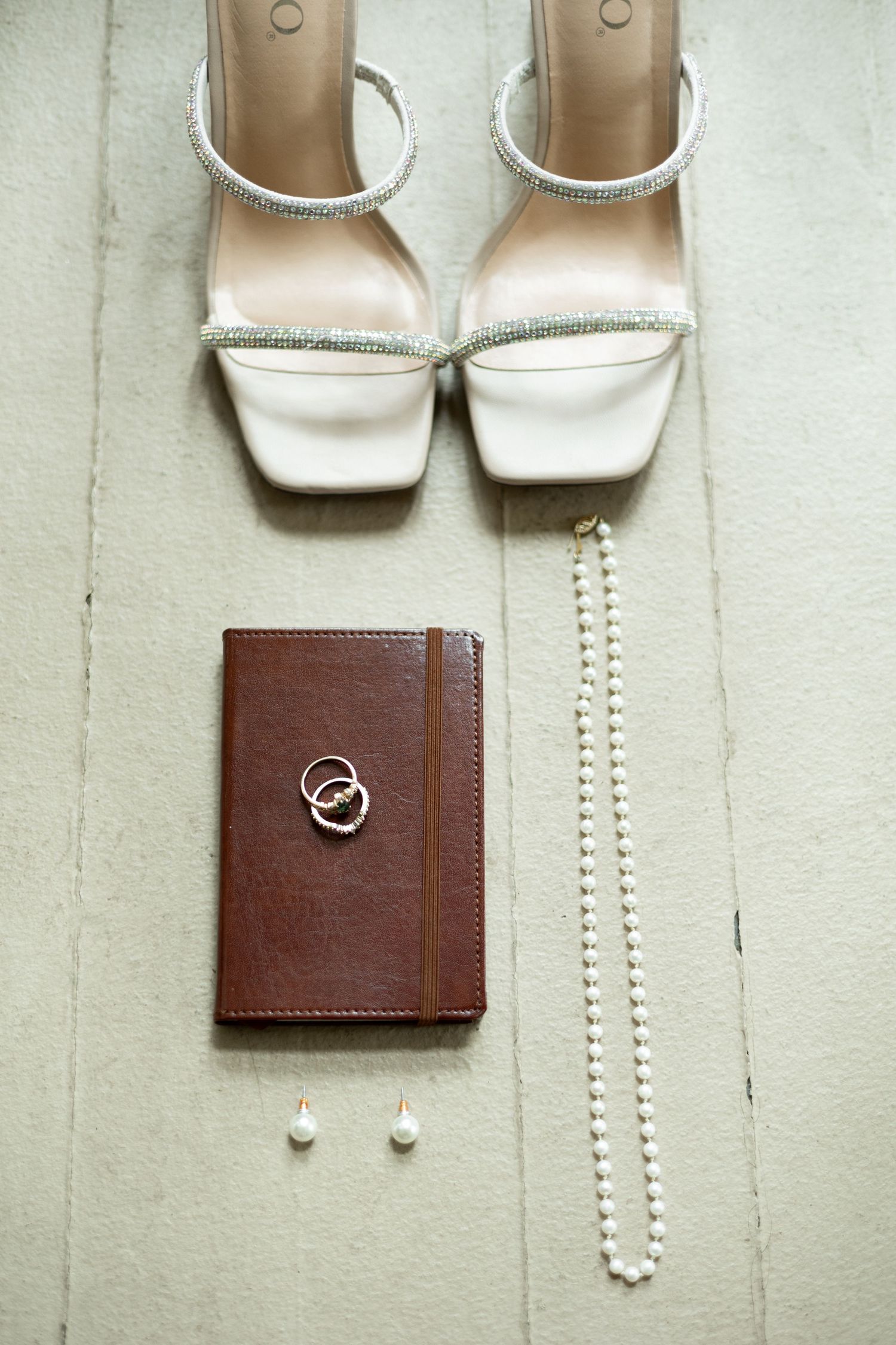 Wedding accessories flatlay with white shoes, brown leather wallet, rings and pearl necklace.