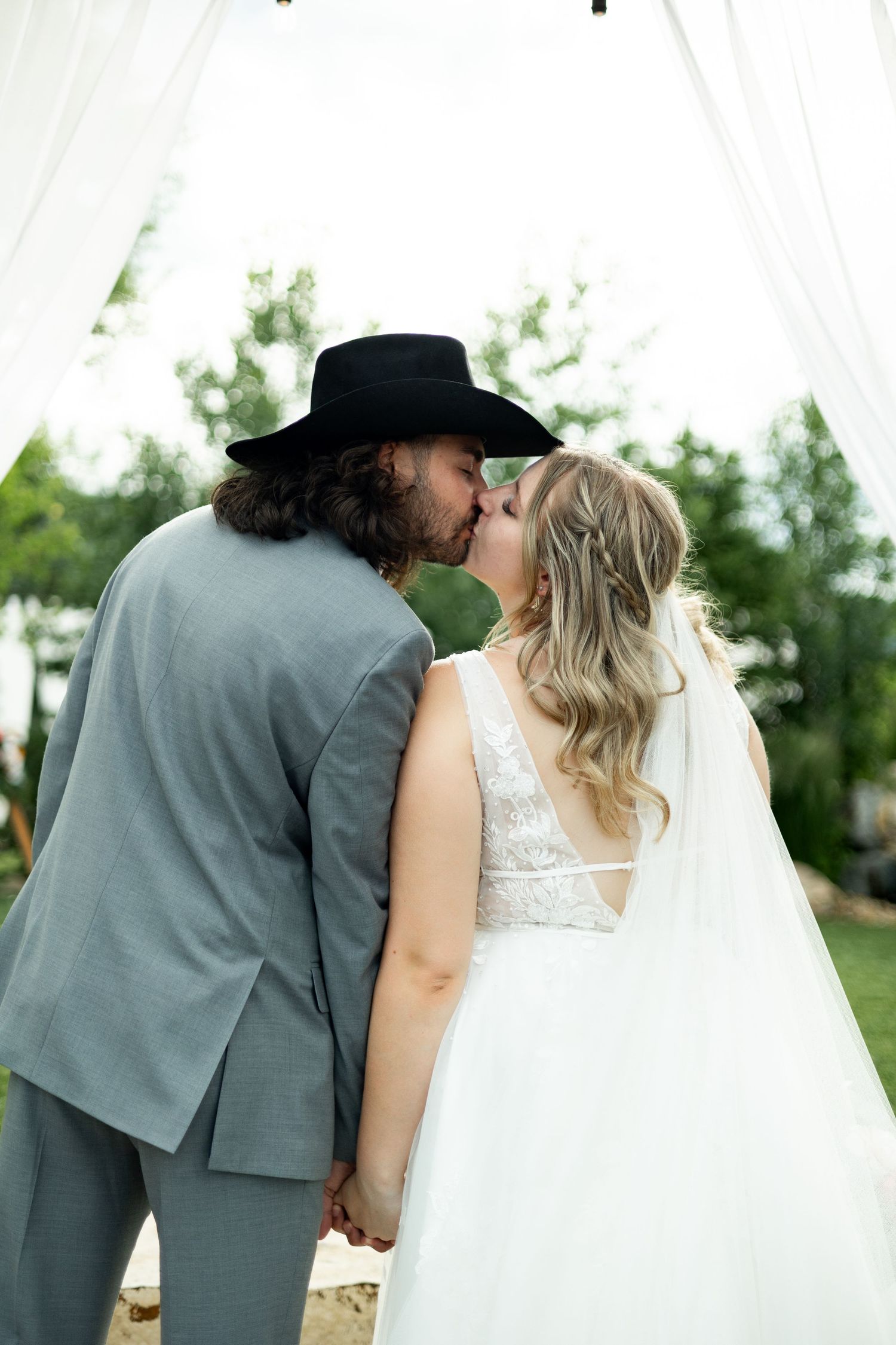 A wedding couple in a grey suit and white dress share a romantic kiss outdoors.