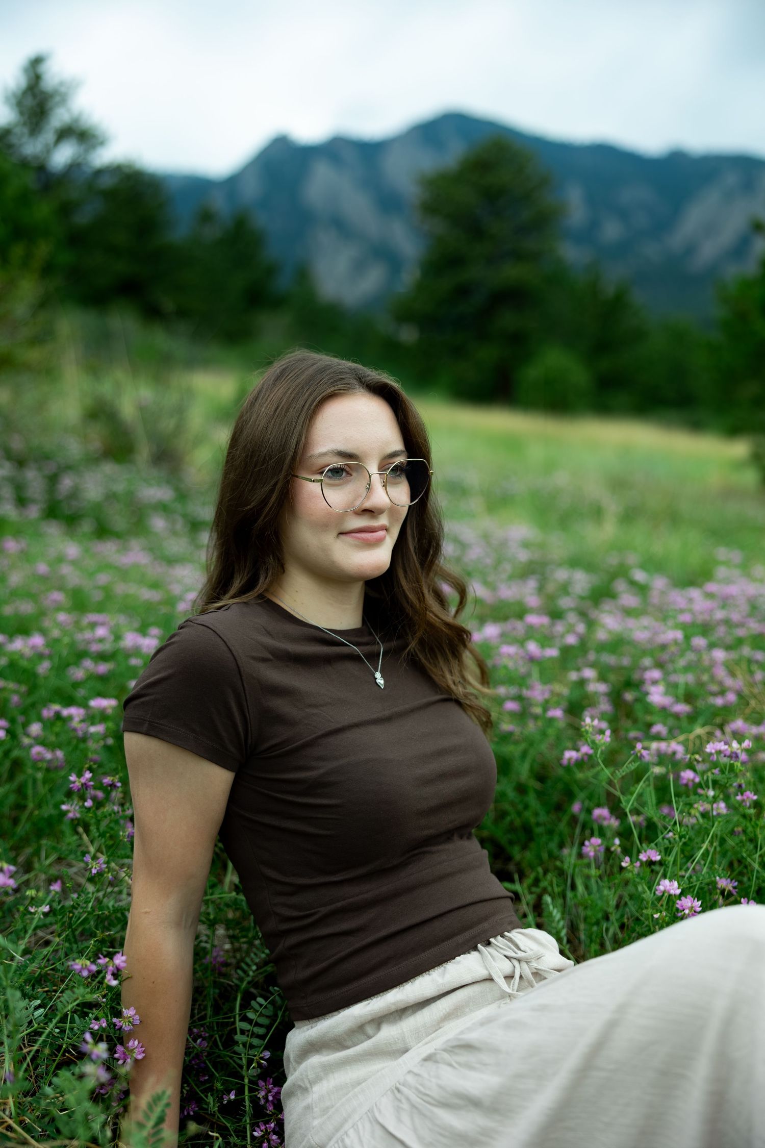Young woman with glasses sitting in a wildflower meadow, wearing a brown top, with mountains in the background.