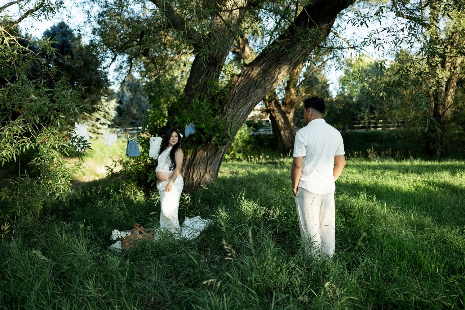 Couple in white outfits standing in lush green grass under a large tree during golden hour outdoors.
