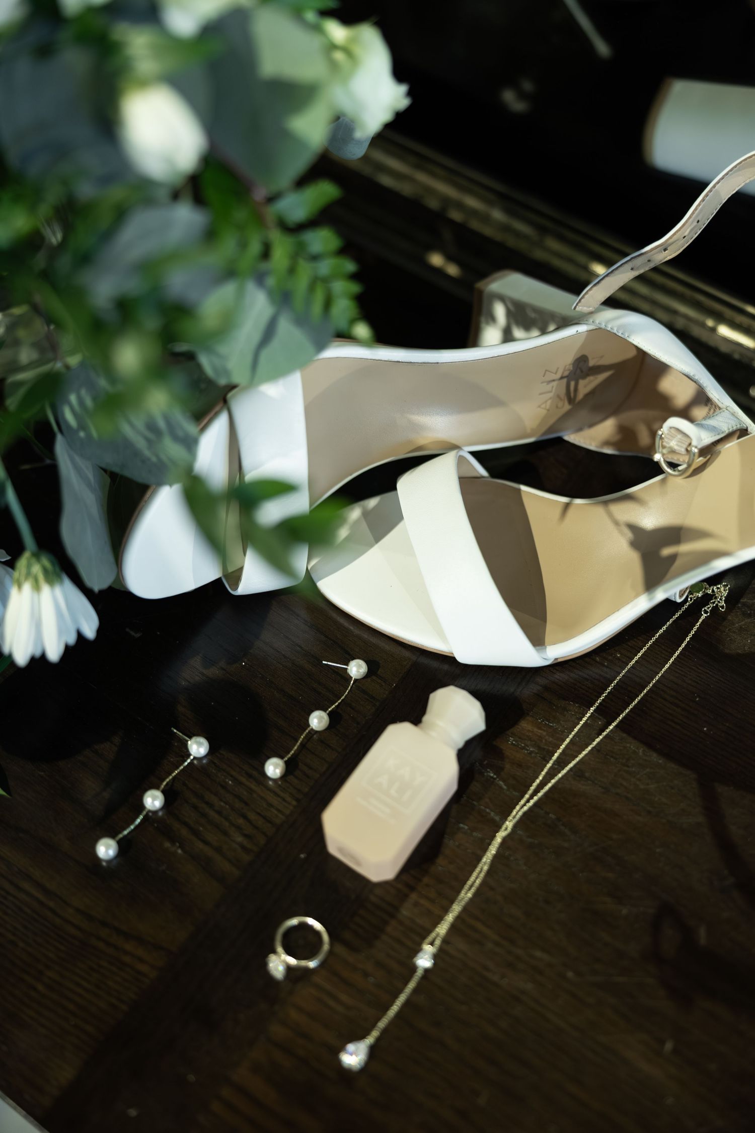 Elegant bridal accessories including white heels, pearl jewelry, a ring, and perfume bottle on a dark wood surface.