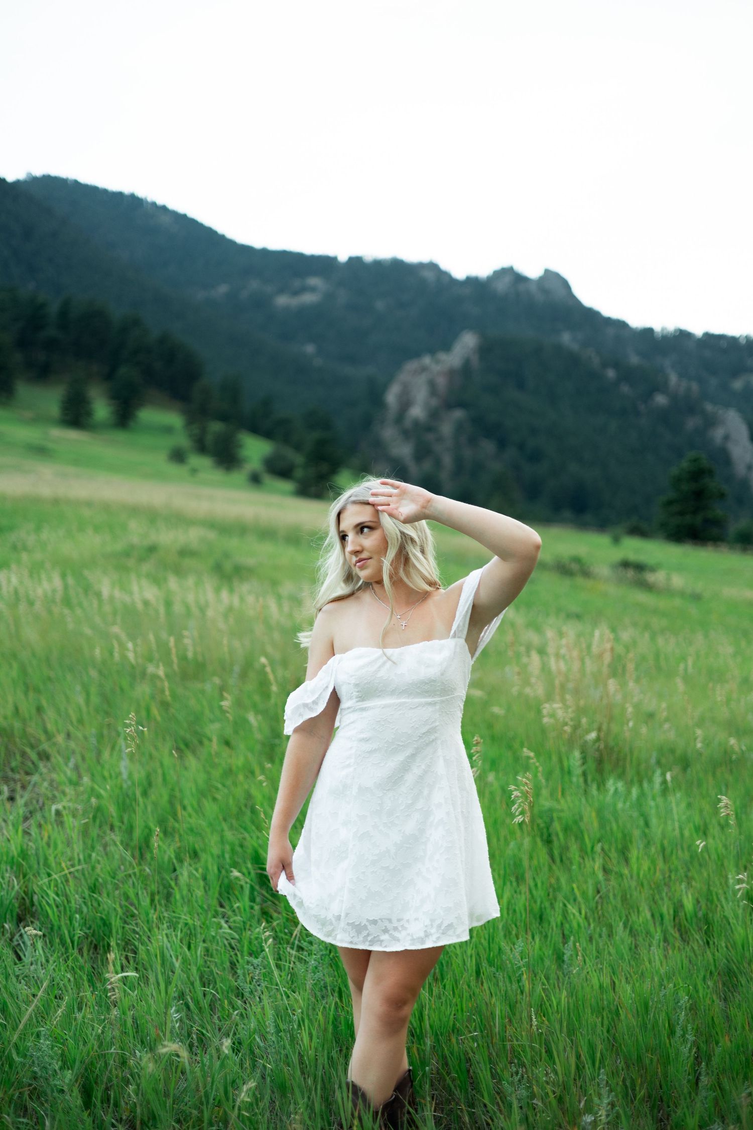 Person in white off-shoulder dress poses in a mountain meadow with green grass and hills in the background.