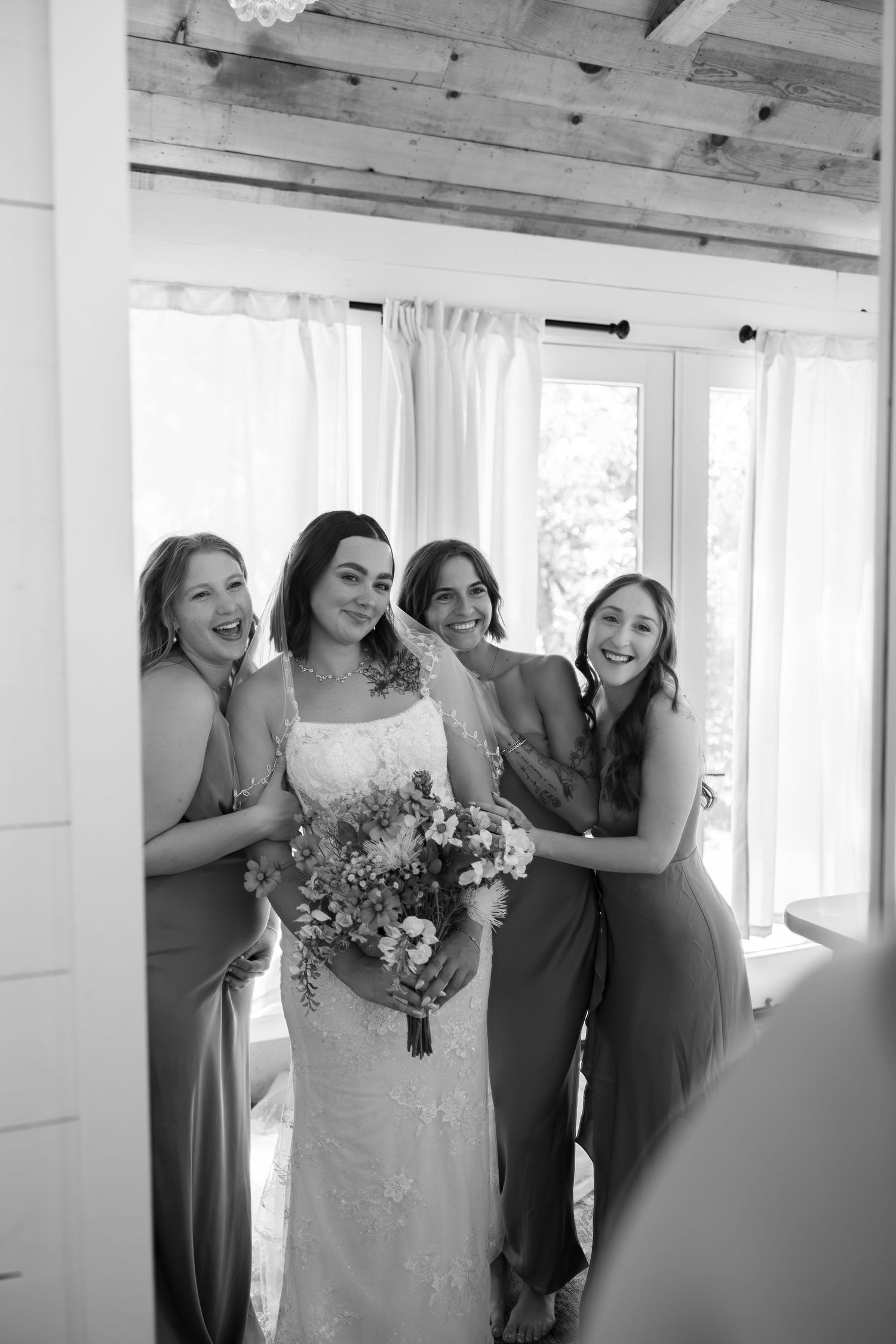 Bride holding bouquet poses with smiling bridesmaids in gowns inside a bright room with sheer curtains.