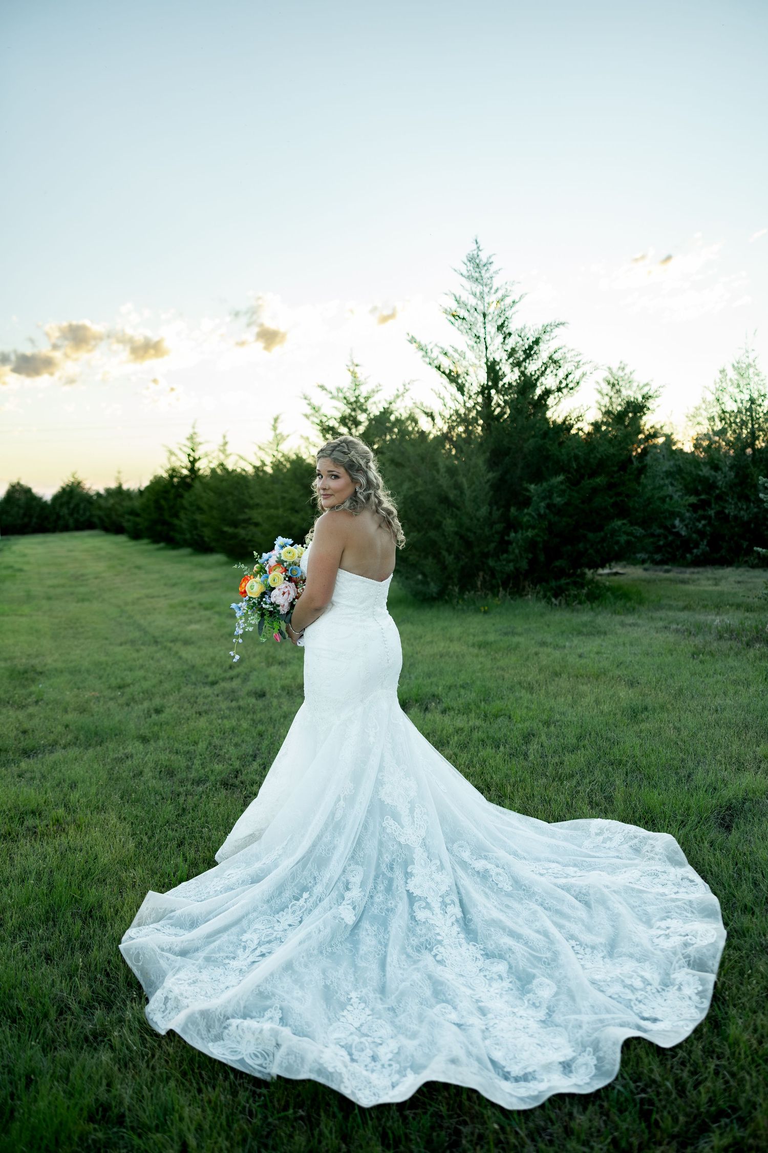 Bride in lace mermaid wedding gown with train holds bouquet outdoors at sunset near evergreen trees.