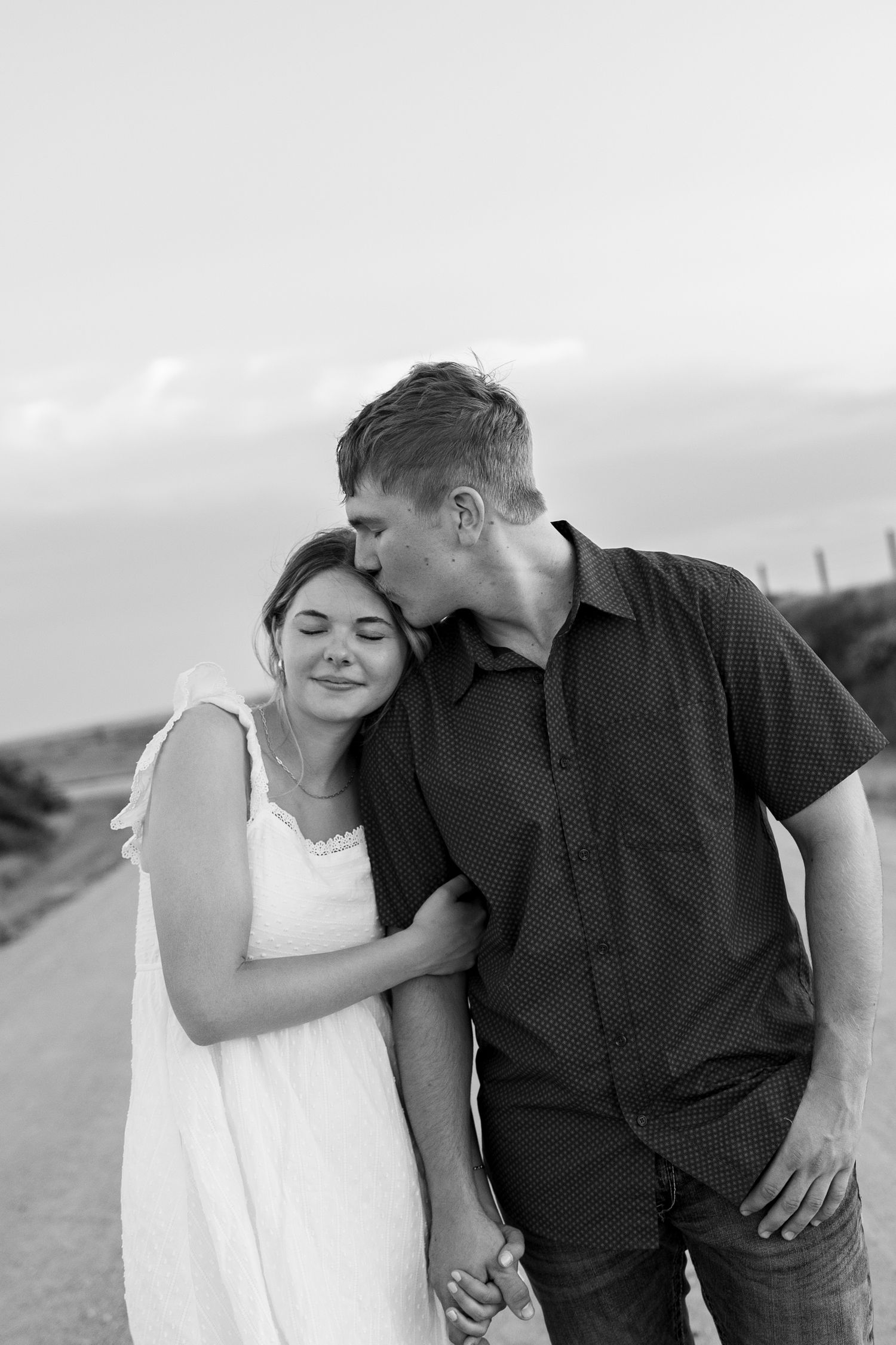 Black and white photo of a couple holding hands on a road, man kissing woman on forehead as she smiles.