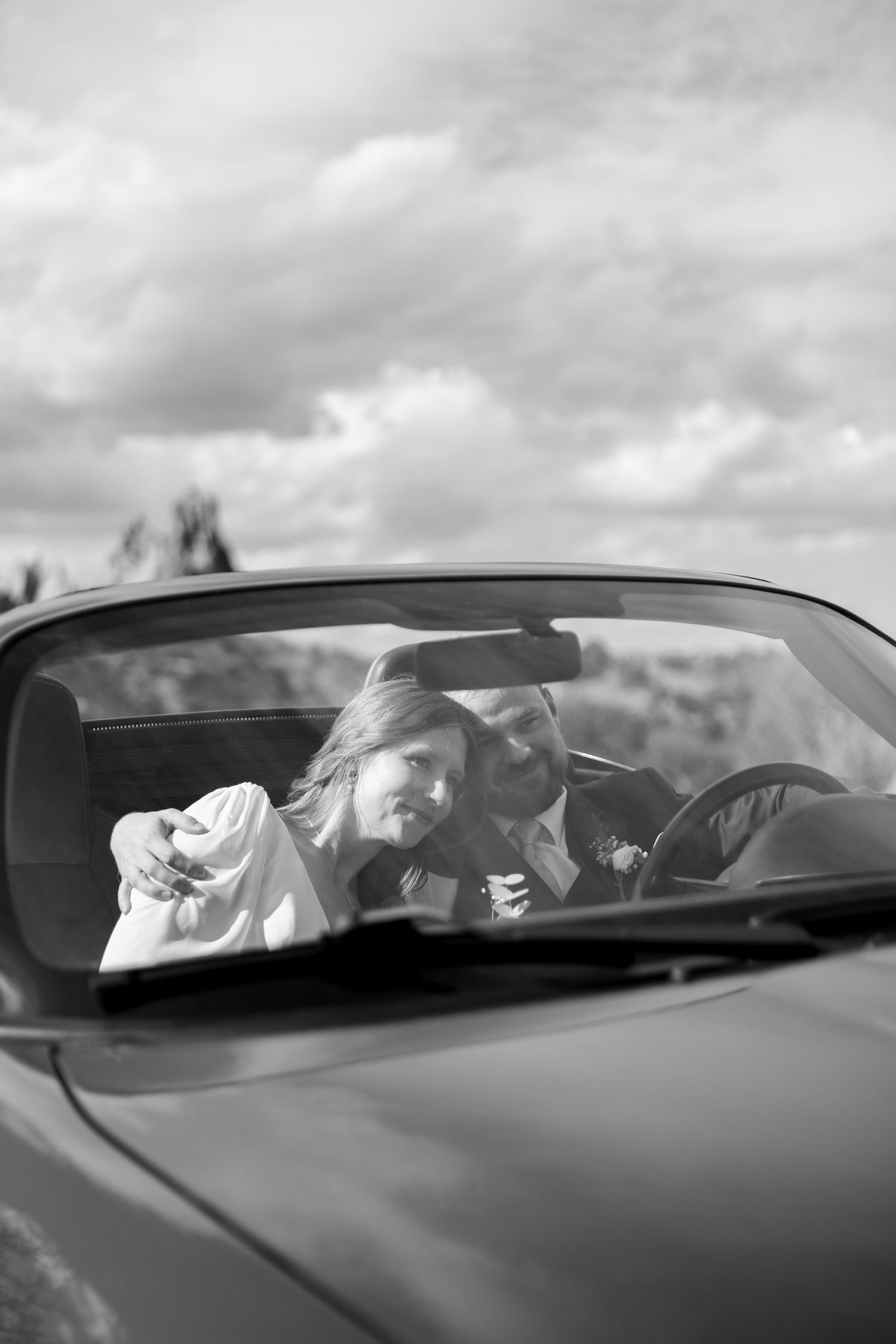 Black and white photo of a person reclining in a convertible car while looking up at cloudy skies.