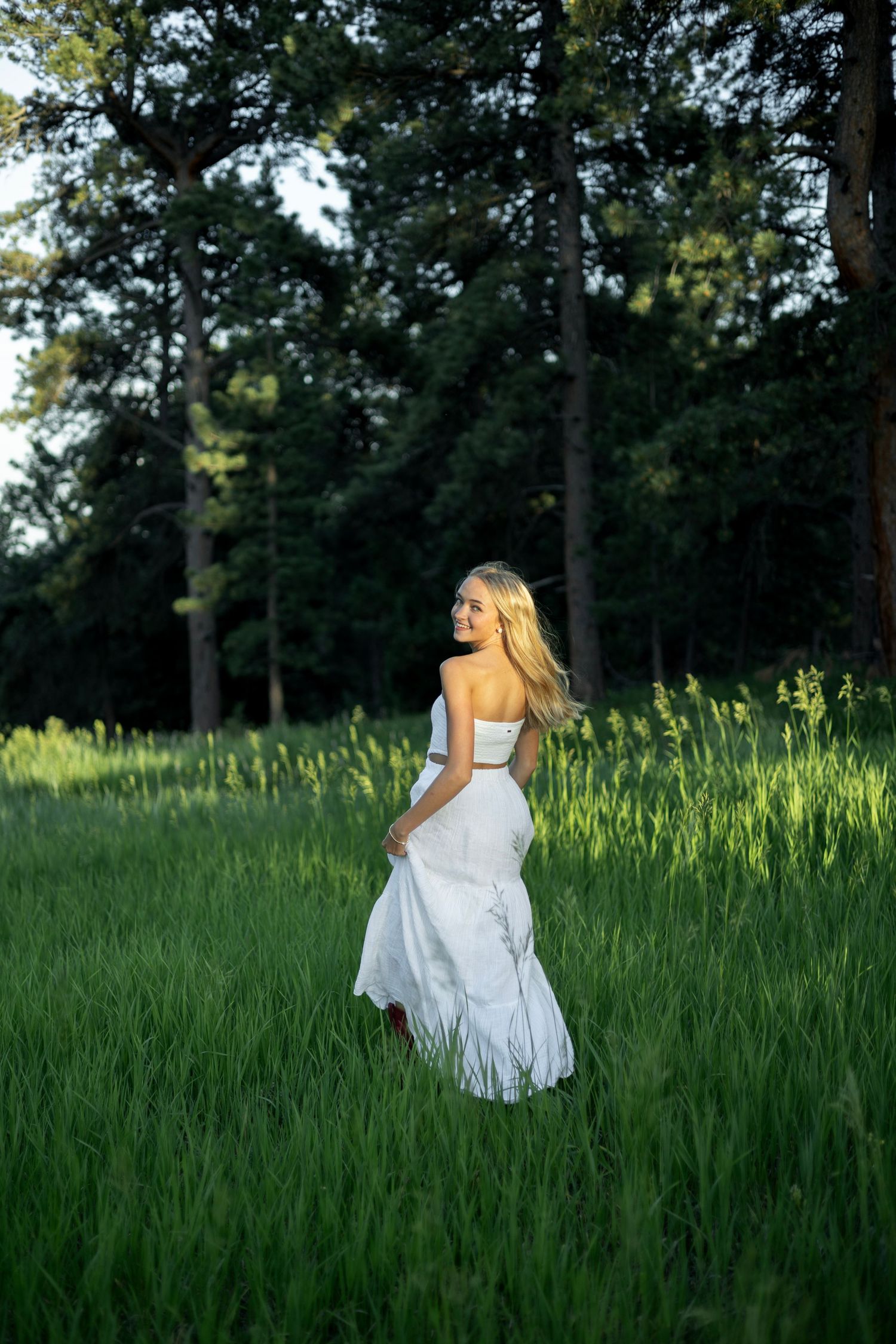 A white flowing dress catches late afternoon light in a meadow surrounded by tall pine trees in the forest.