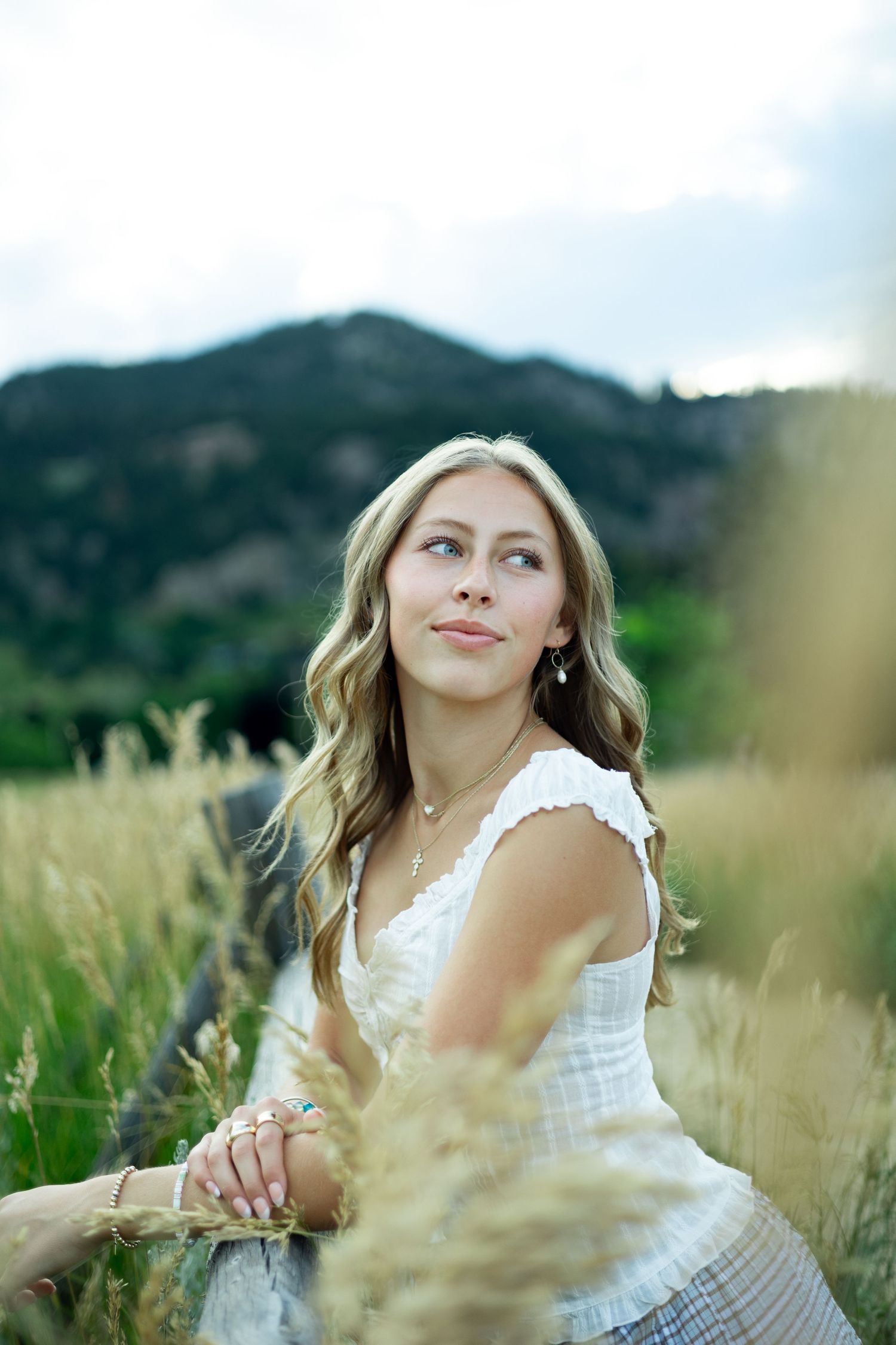 Person in white dress sits gracefully among tall grass with mountain backdrop on sunny afternoon.
