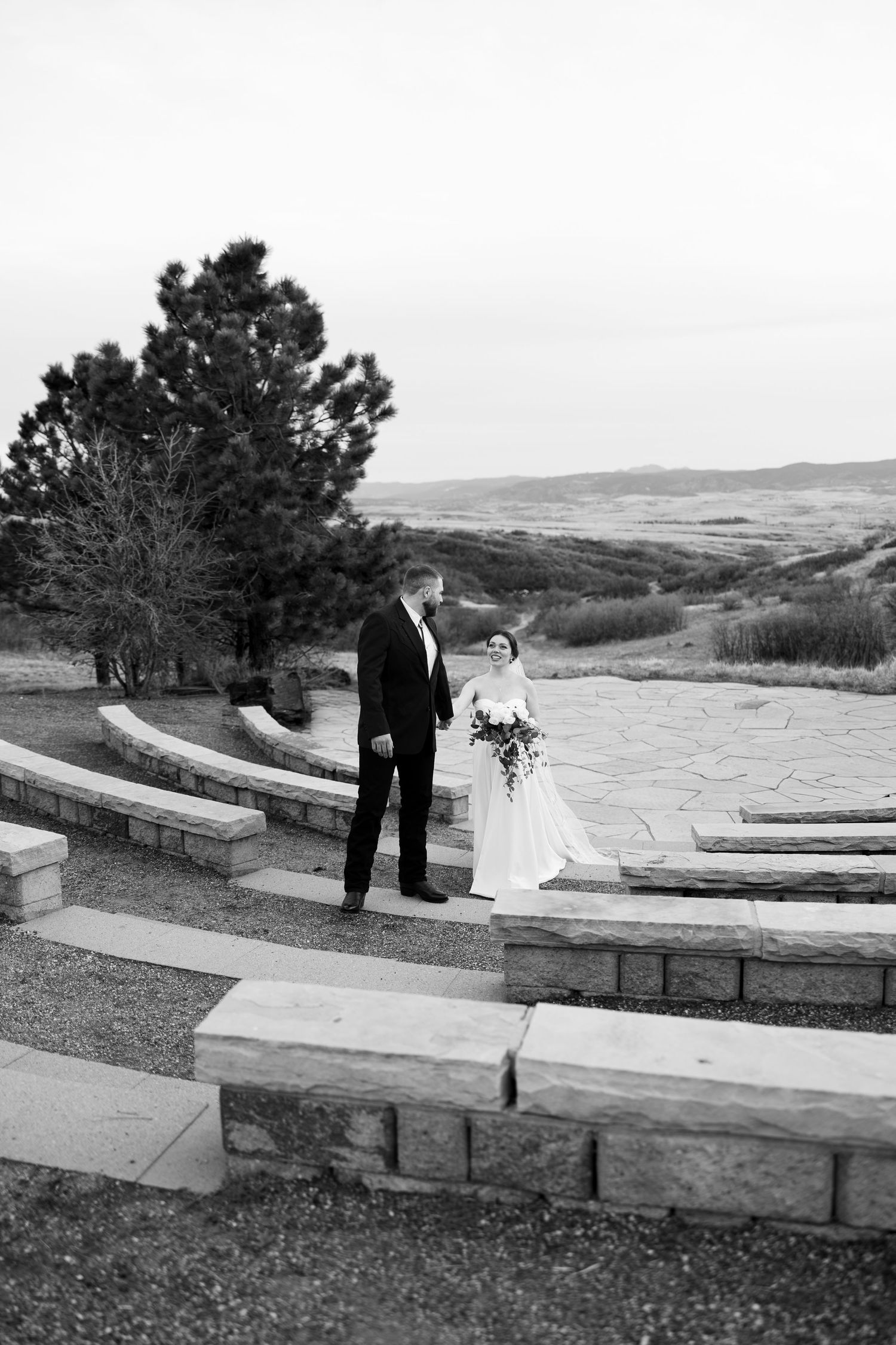 Black and white photo of bride and groom standing on stone steps at an outdoor wedding venue with scenic landscape views.