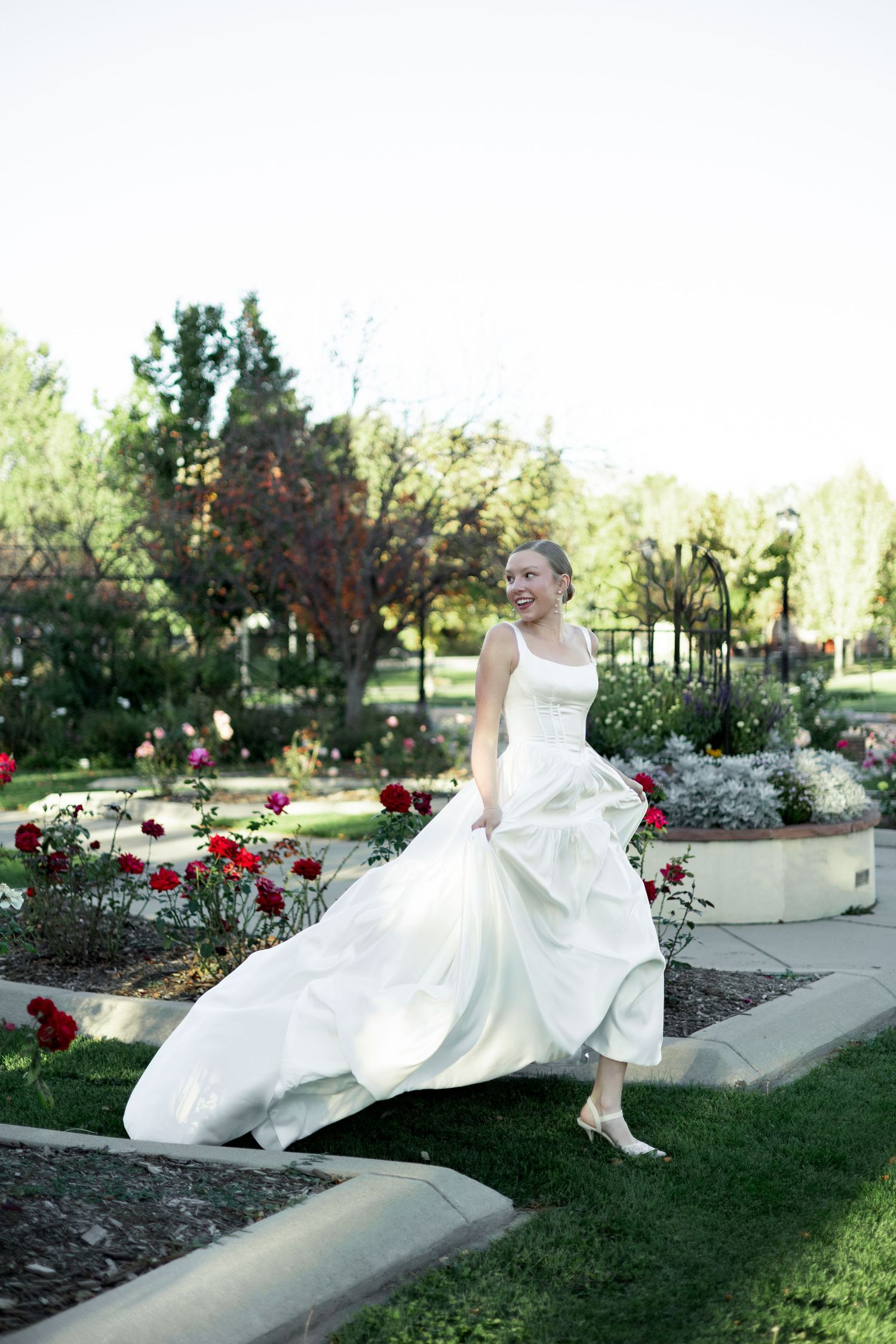 A white wedding dress flows elegantly across a garden wall as someone poses gracefully in an outdoor setting with red roses.