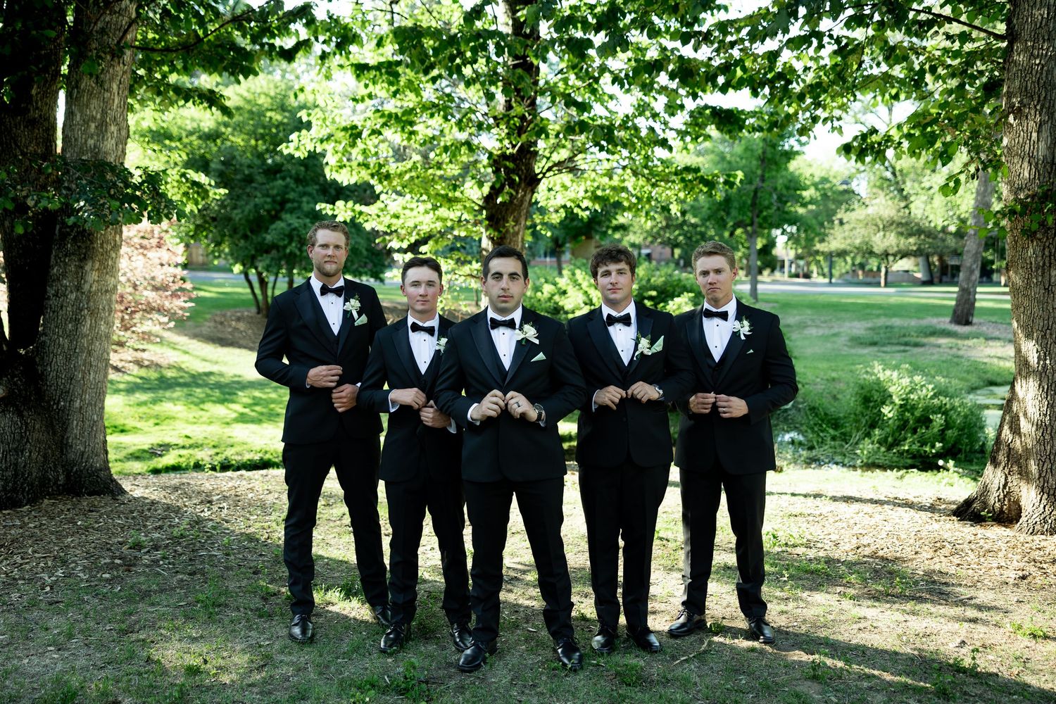 Five groomsmen in black tuxedos with bow ties and boutonnieres pose outdoors under green trees at a wedding.