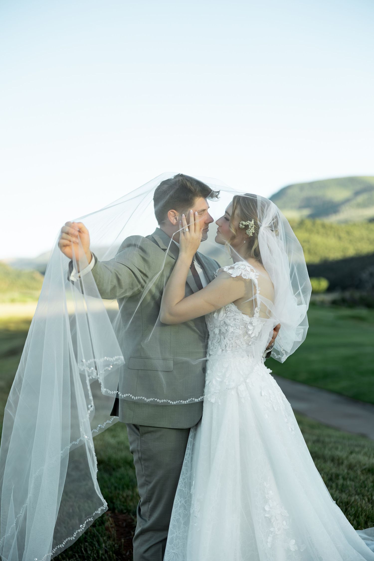 Bride and groom share romantic moment under flowing white veil with scenic mountain backdrop on their wedding day.