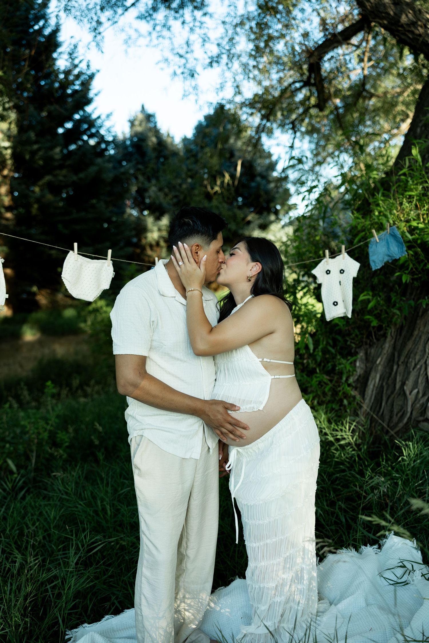 Pregnant couple in white outfits kissing outdoors with baby clothes hanging on a line in a lush green garden.