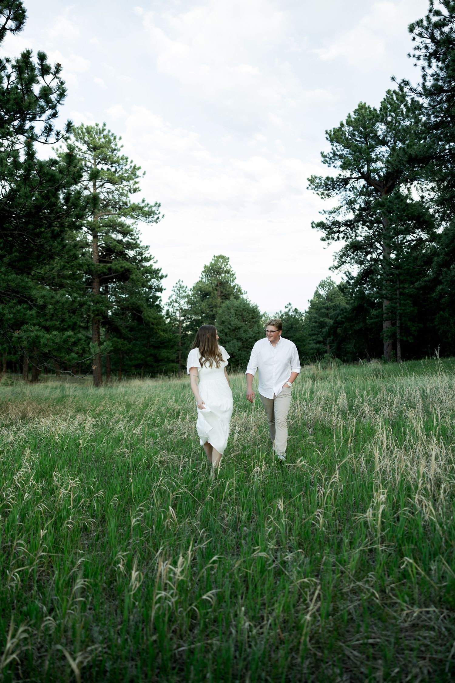 Couple in white outfits holding hands while walking through a lush green meadow surrounded by tall pine trees.