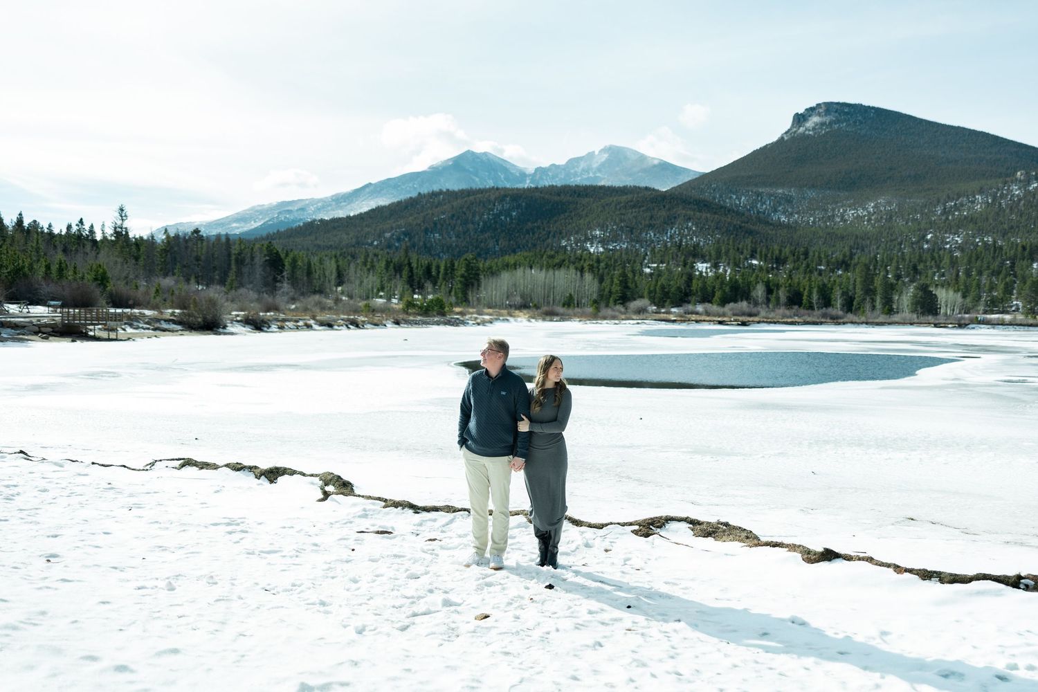 Two people stand together on a snowy shoreline with mountains and forest in the background at a winter lake.