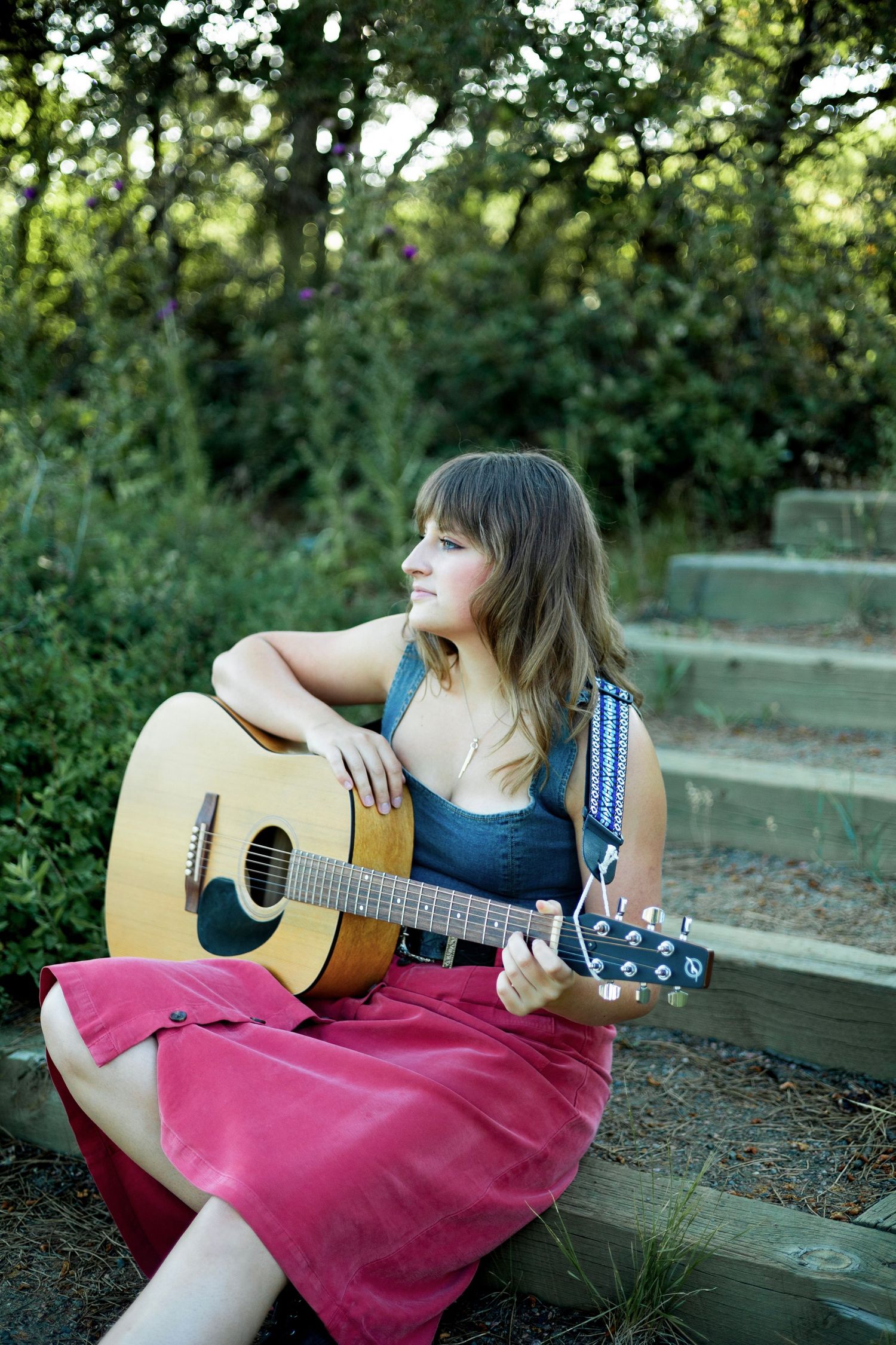Young woman with bangs sits outdoors on wooden steps holding an acoustic guitar, wearing a red skirt and blue top.