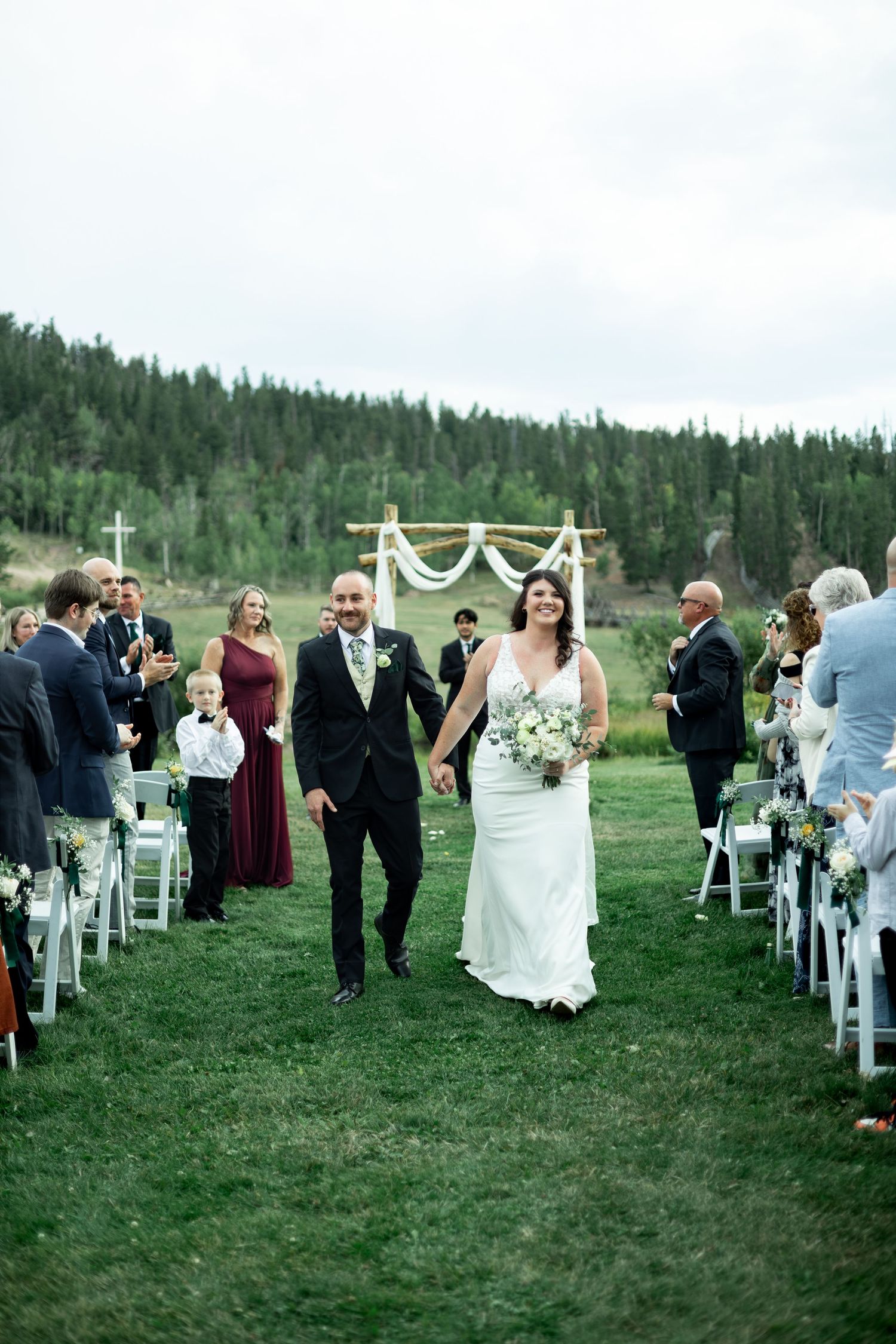 Bride and groom walk down outdoor aisle after wedding ceremony with forest backdrop and guests celebrating.