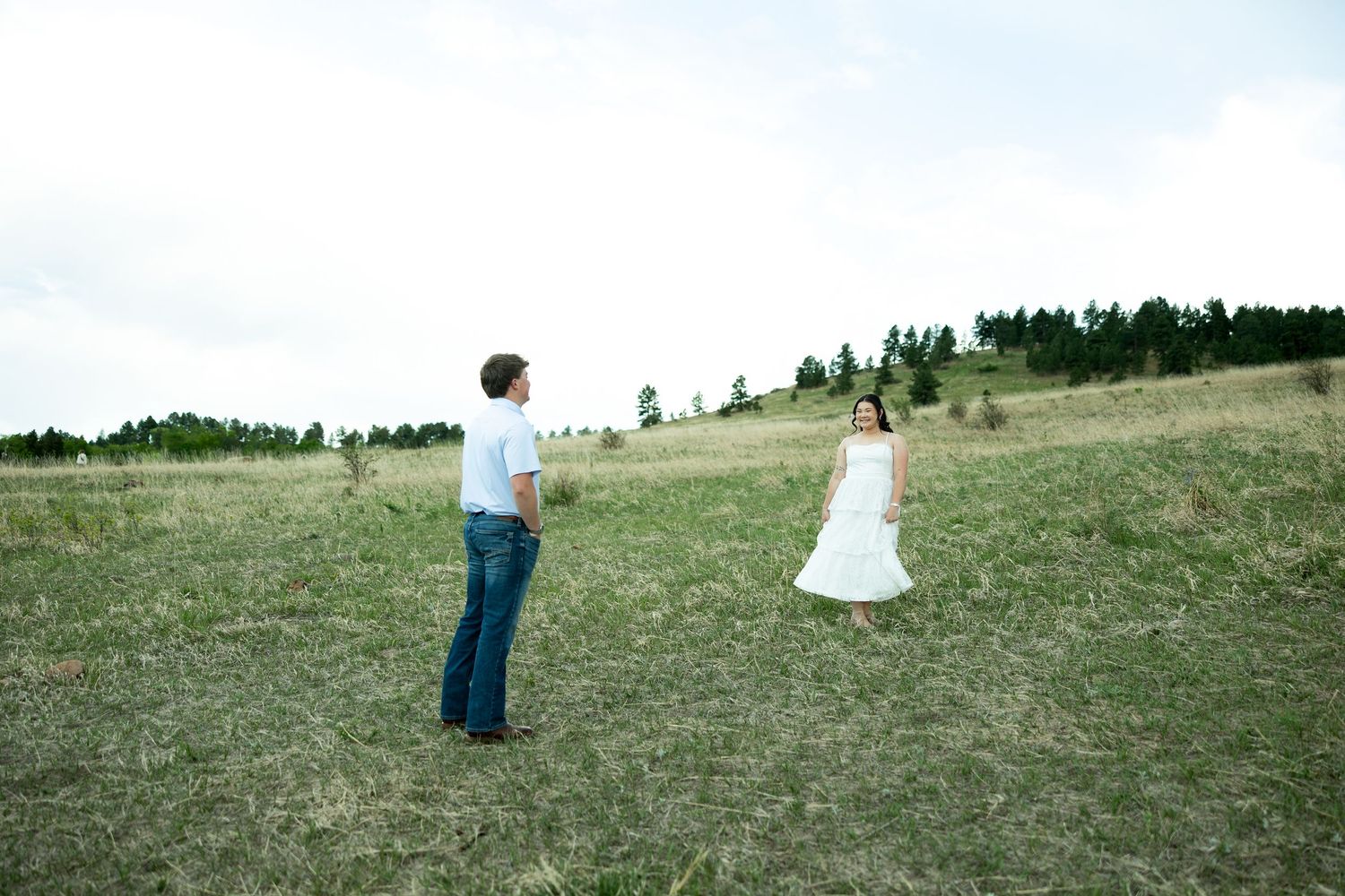 Couple in a green meadow, man in jeans and woman in white dress, with trees on a hill in the background.