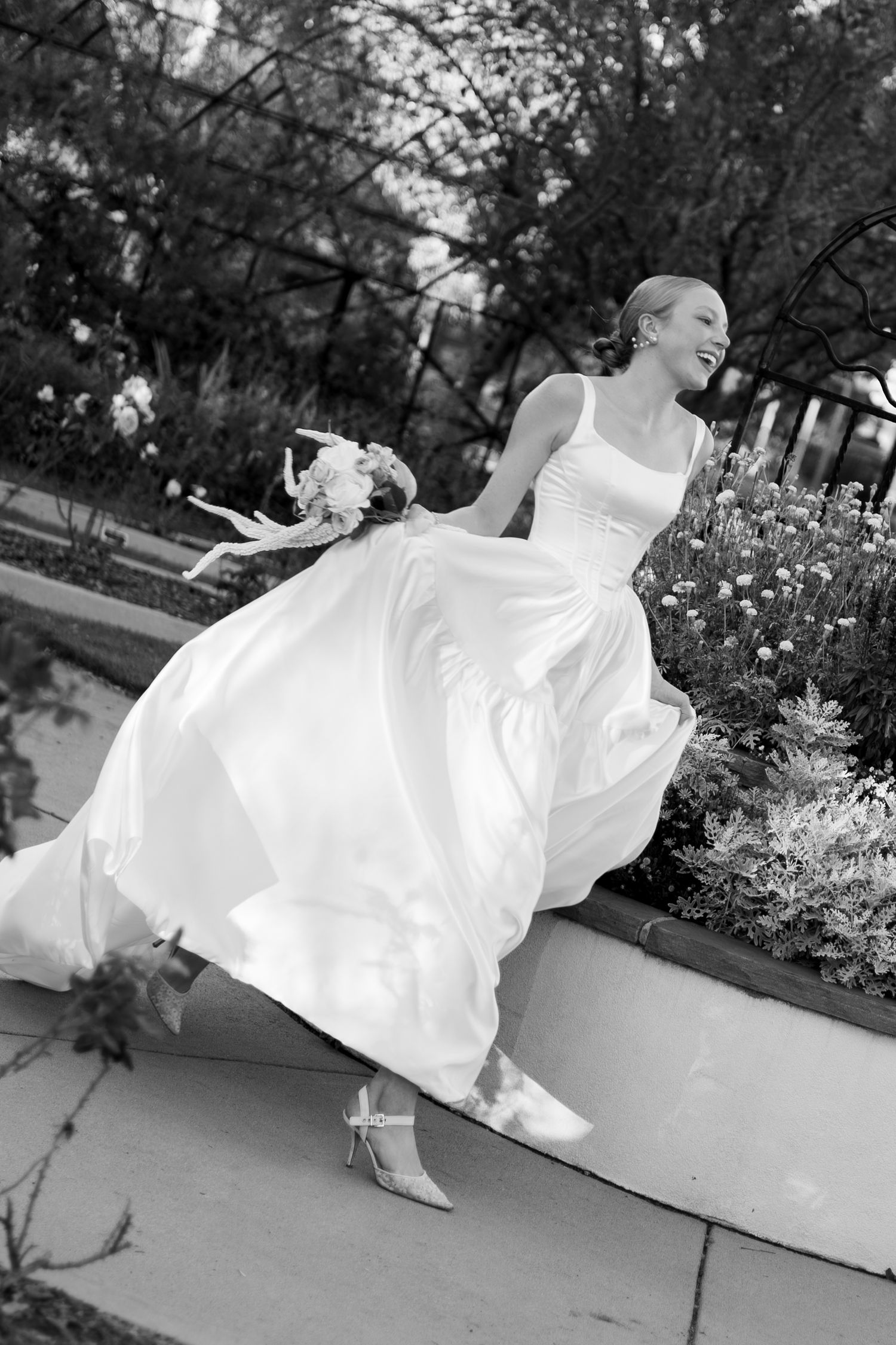 Bride in flowing white gown holding bouquet, laughing joyfully outdoors near garden in black and white photo.