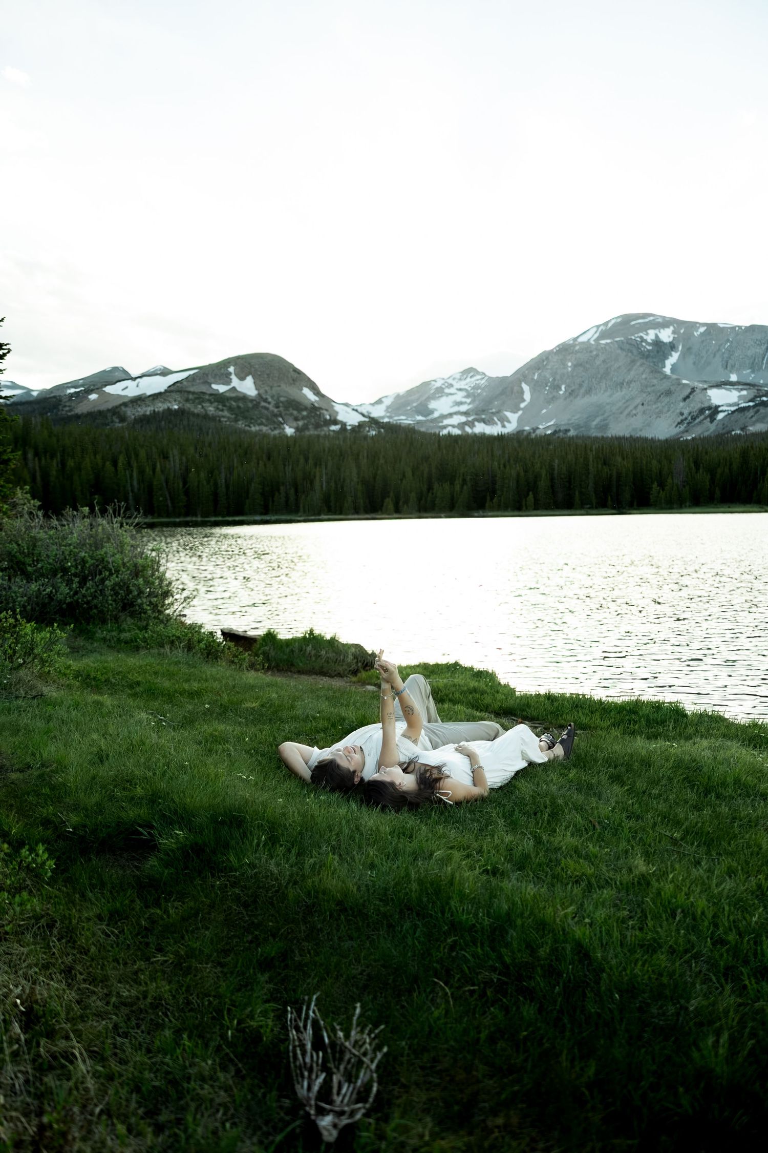 Couple lying on green grass by a serene mountain lake with snow-capped peaks and pine forest backdrop.