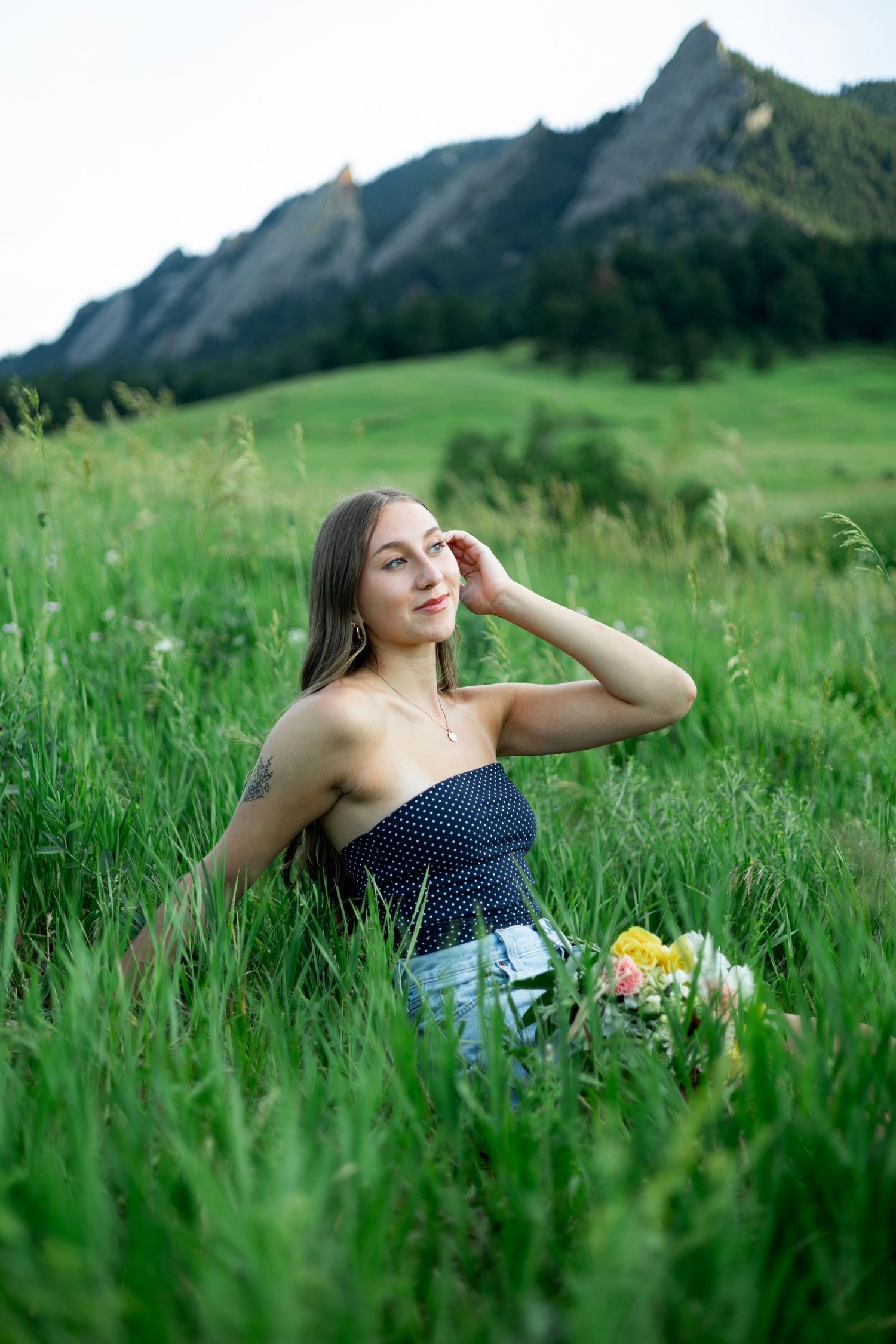 Person in polka dot top sitting in lush mountain meadow with wildflowers and dramatic peaks in background.