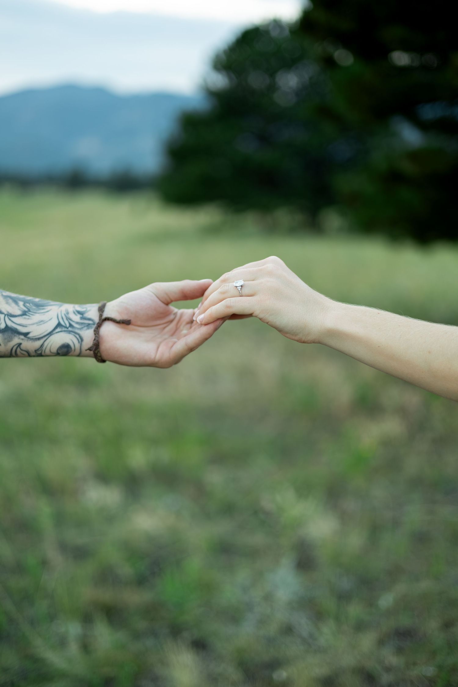 Two hands reach for each other while displaying an engagement ring against a mountain backdrop.