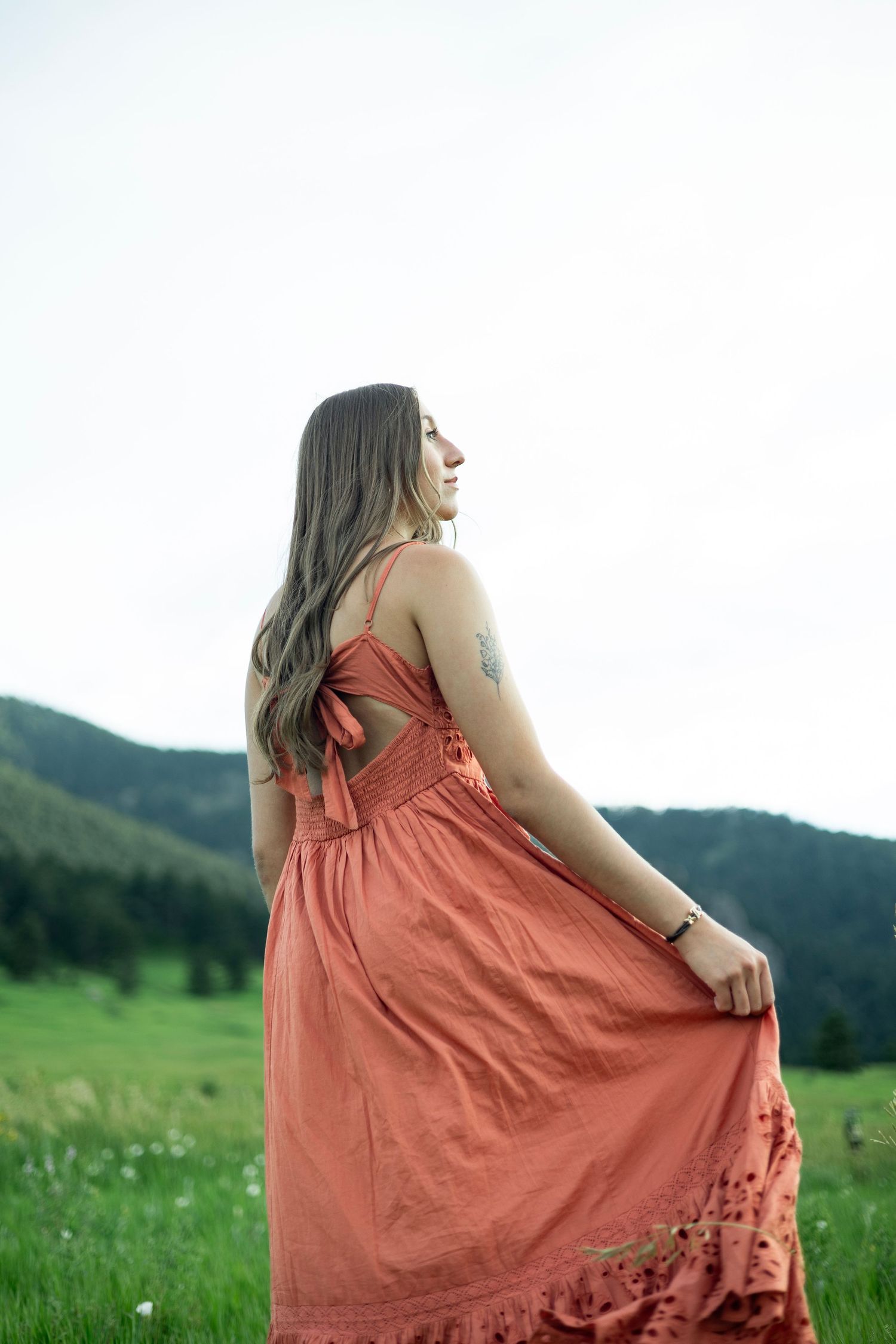 A person in a flowing coral-colored dress stands in a green meadow with mountains in the background.