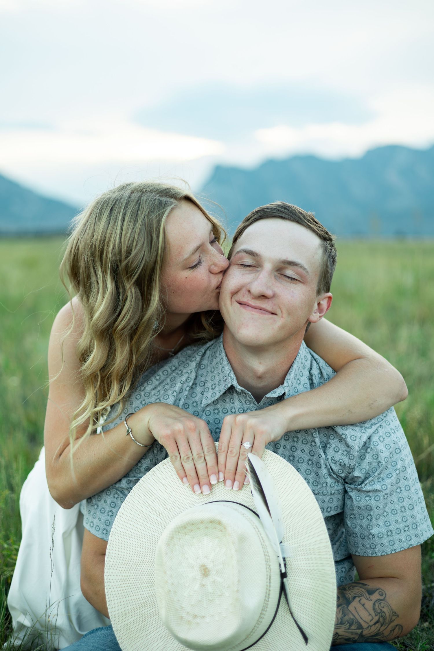 Romantic couple embraces in a grassy field against mountain backdrop while sharing sweet moments with a white hat.