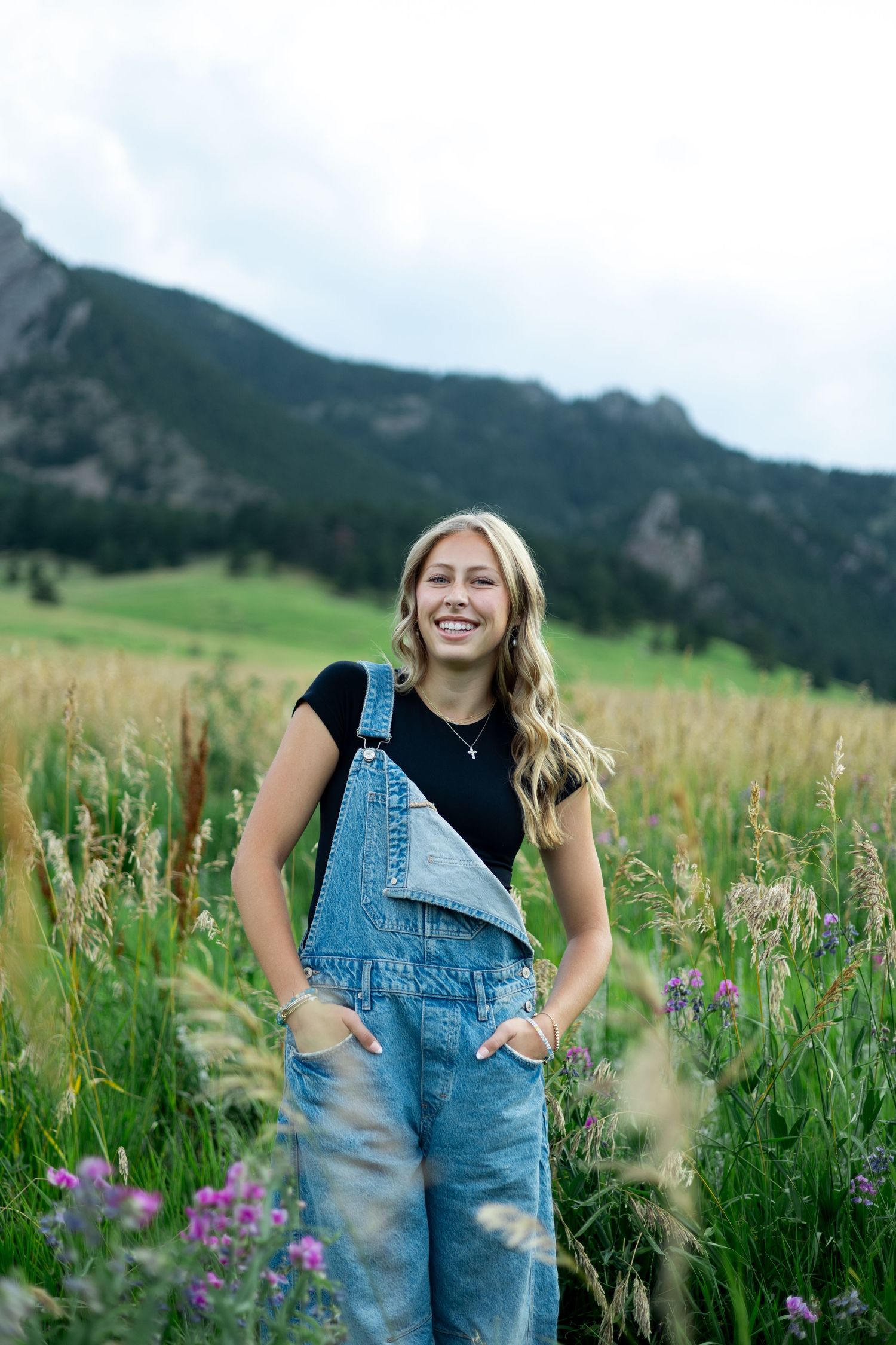 Person in denim overalls and black shirt smiling in a mountain meadow with purple wildflowers and green grass.