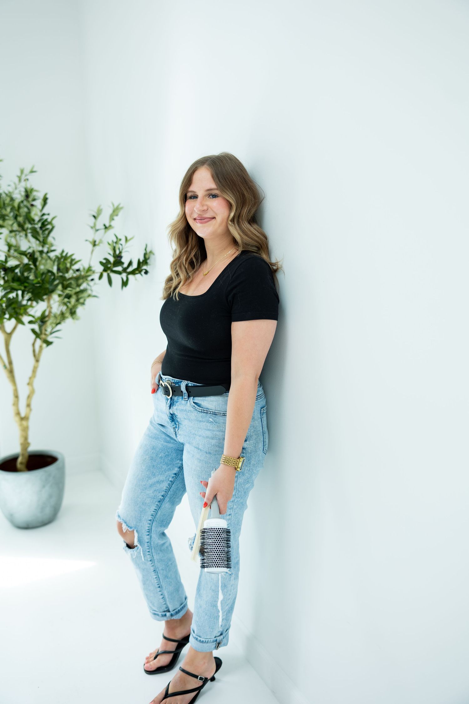 Woman in black top and ripped jeans leaning against white wall, smiling, with potted plant nearby.