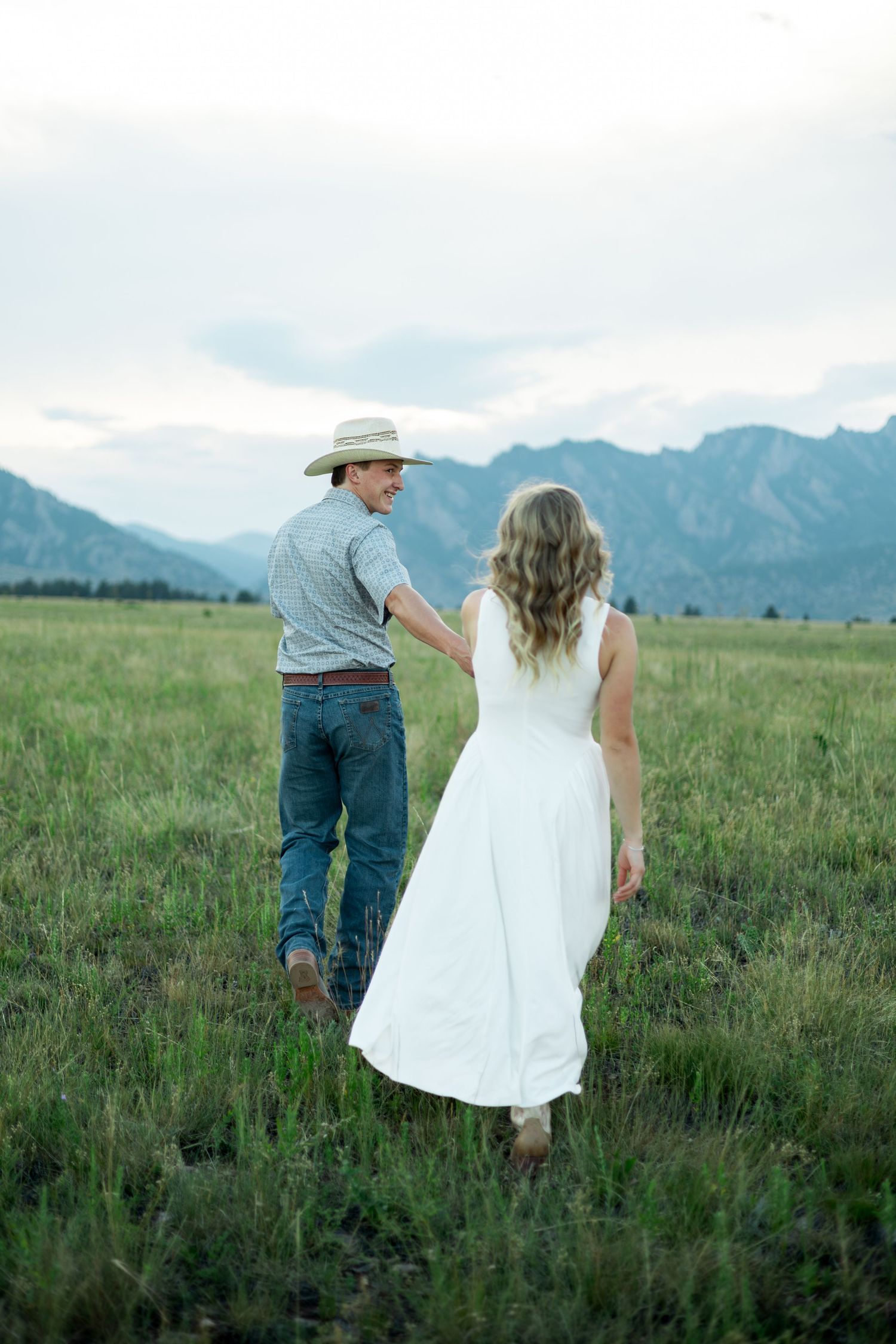Couple holding hands walking through green mountain meadow, man in cowboy hat and jeans, woman in white dress.