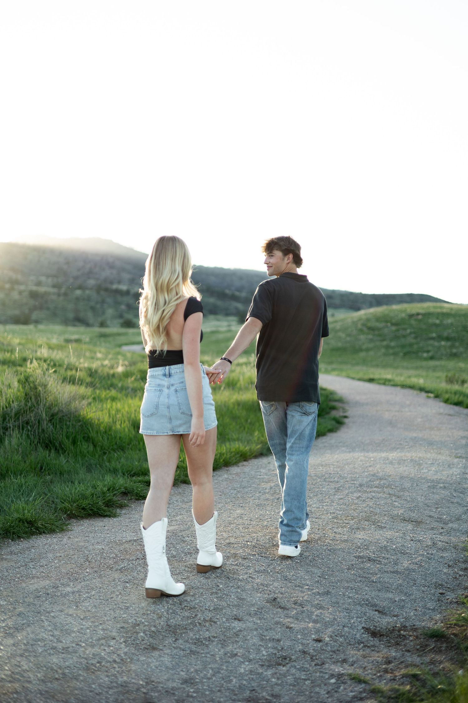 Couple holding hands walking down a winding path through green hills at sunset, woman wearing denim skirt and white cowboy boots.