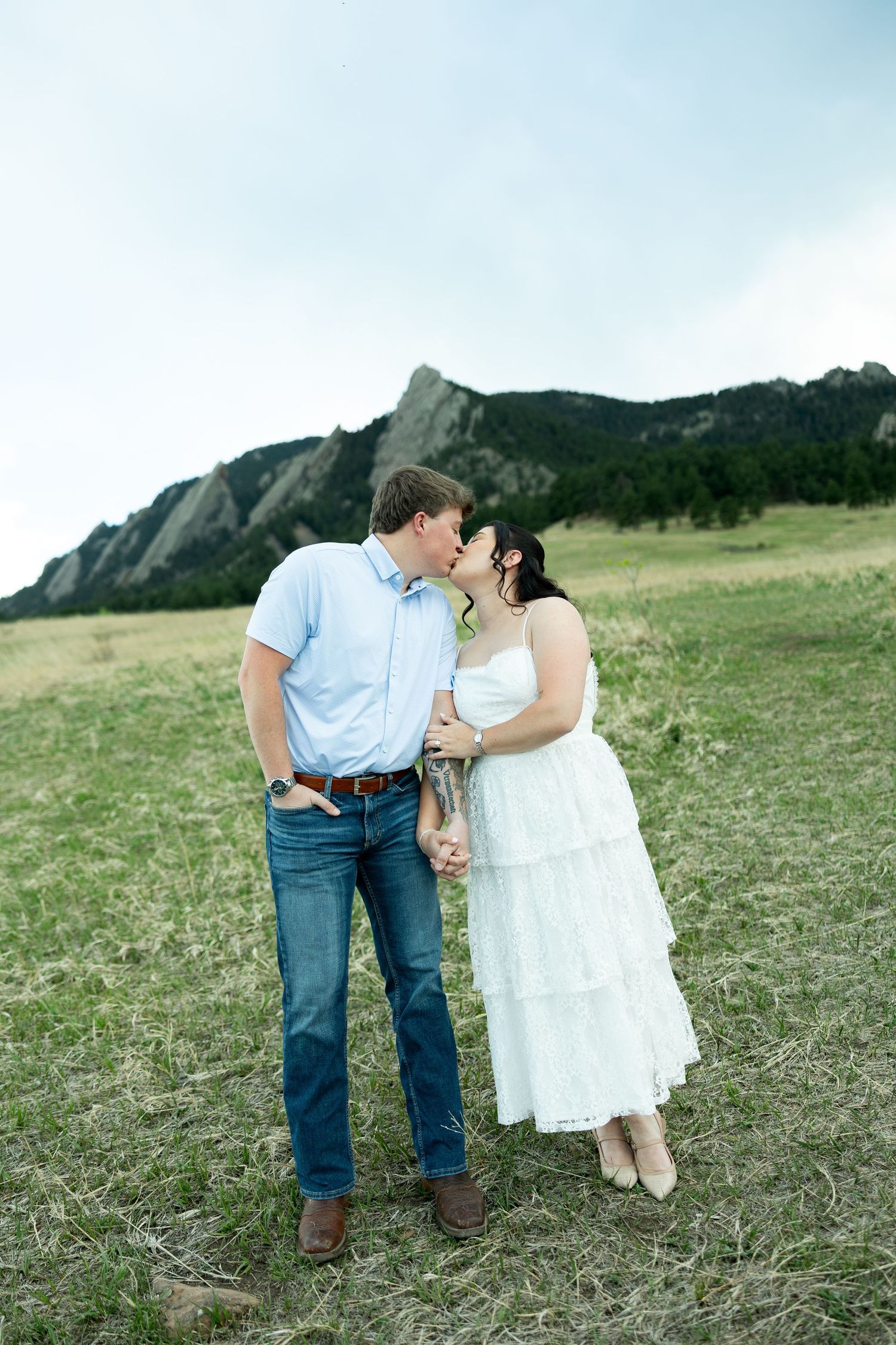 Couple sharing a kiss in a grassy field with the iconic Flatirons rock formations in Boulder, Colorado behind them.