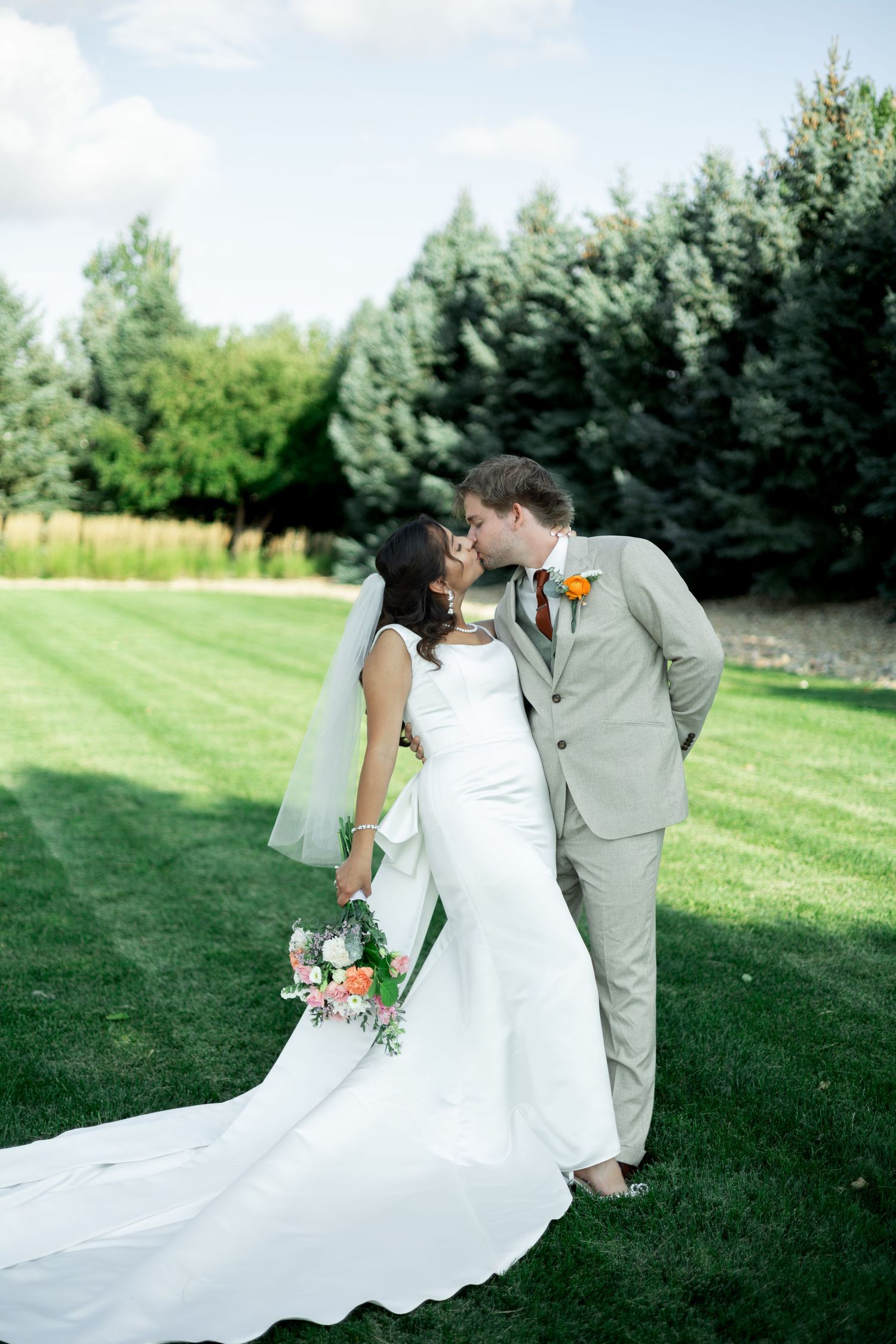 Bride and groom share a romantic kiss on lush green lawn, bride holding colorful bouquet in elegant white gown with train.