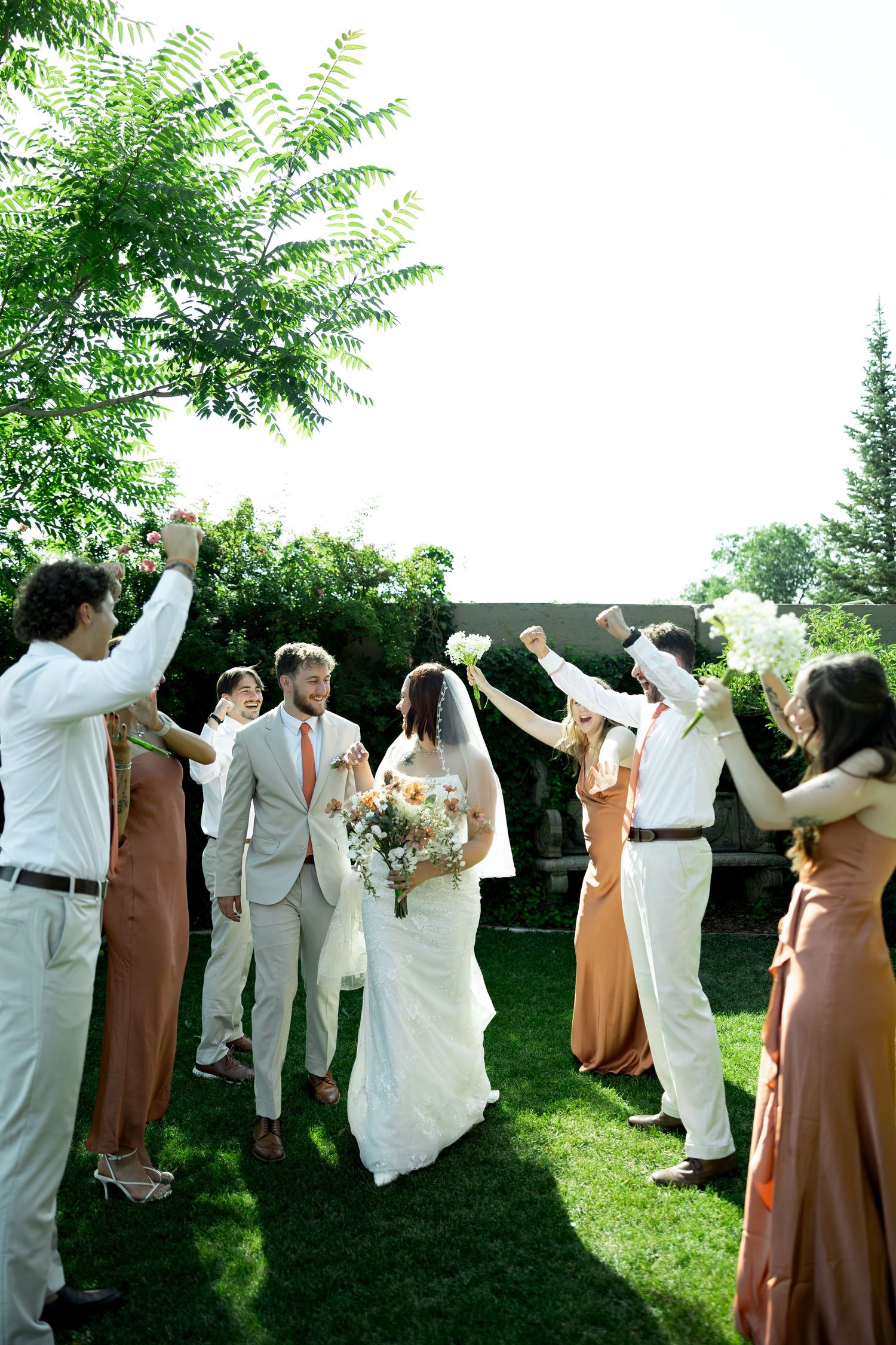 Bride and groom walk through cheering guests in garden wedding ceremony, bridesmaids in rust dresses raise hands.