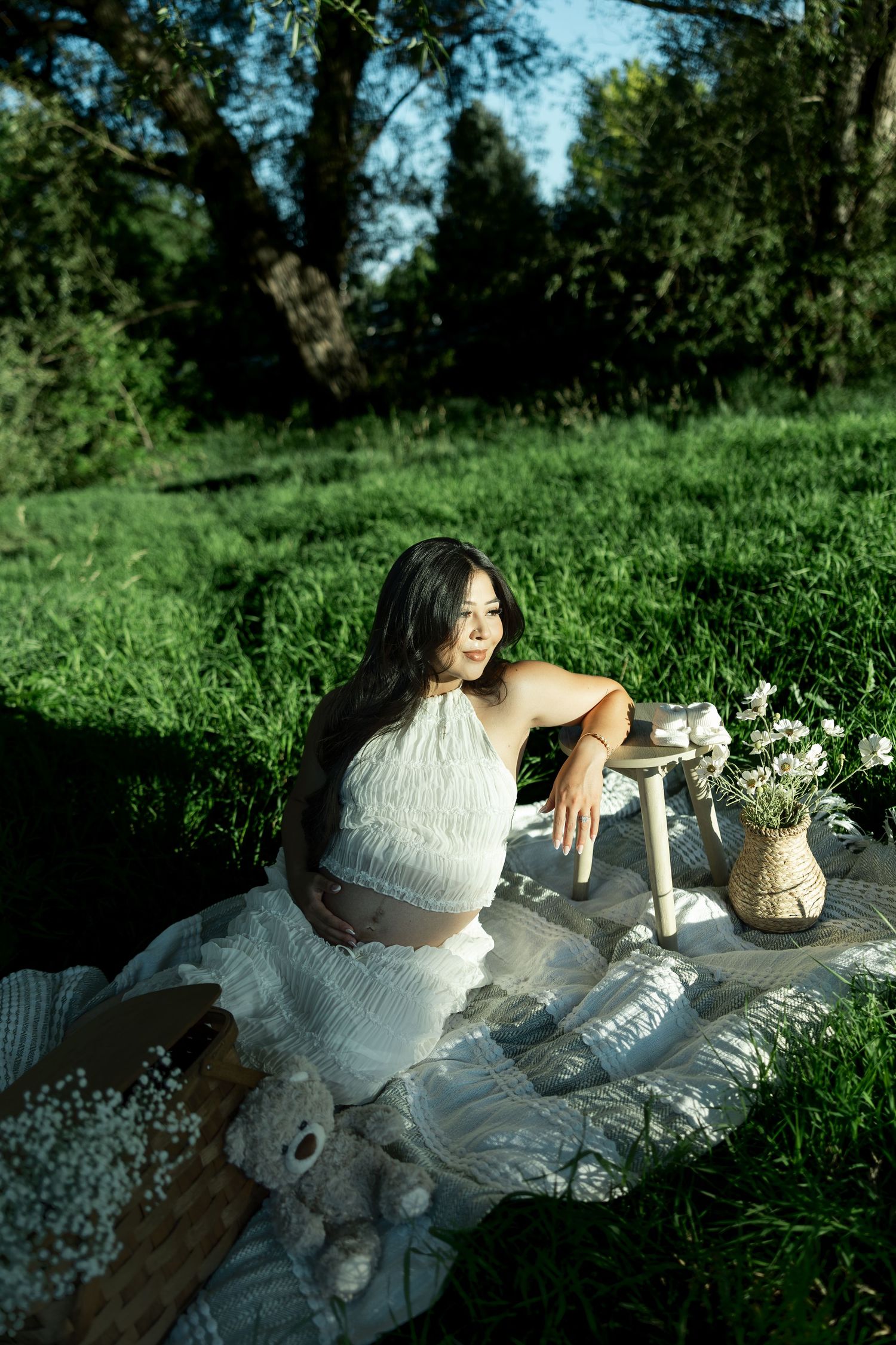 Pregnant woman in white fringe outfit sits on blanket in lush green park with flowers and teddy bear.