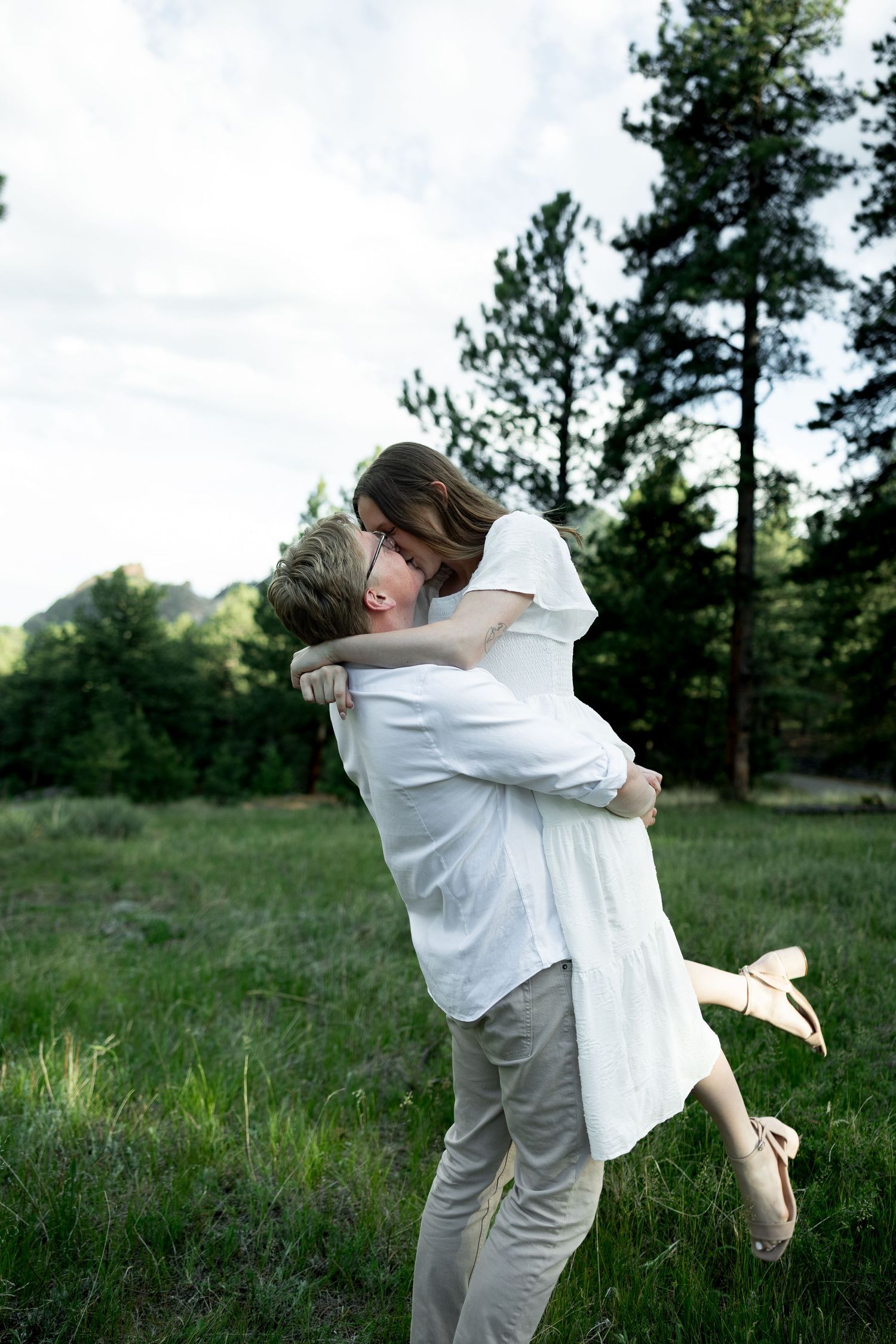 Couple embracing in a green meadow, man lifting woman in white dress, surrounded by tall pine trees.