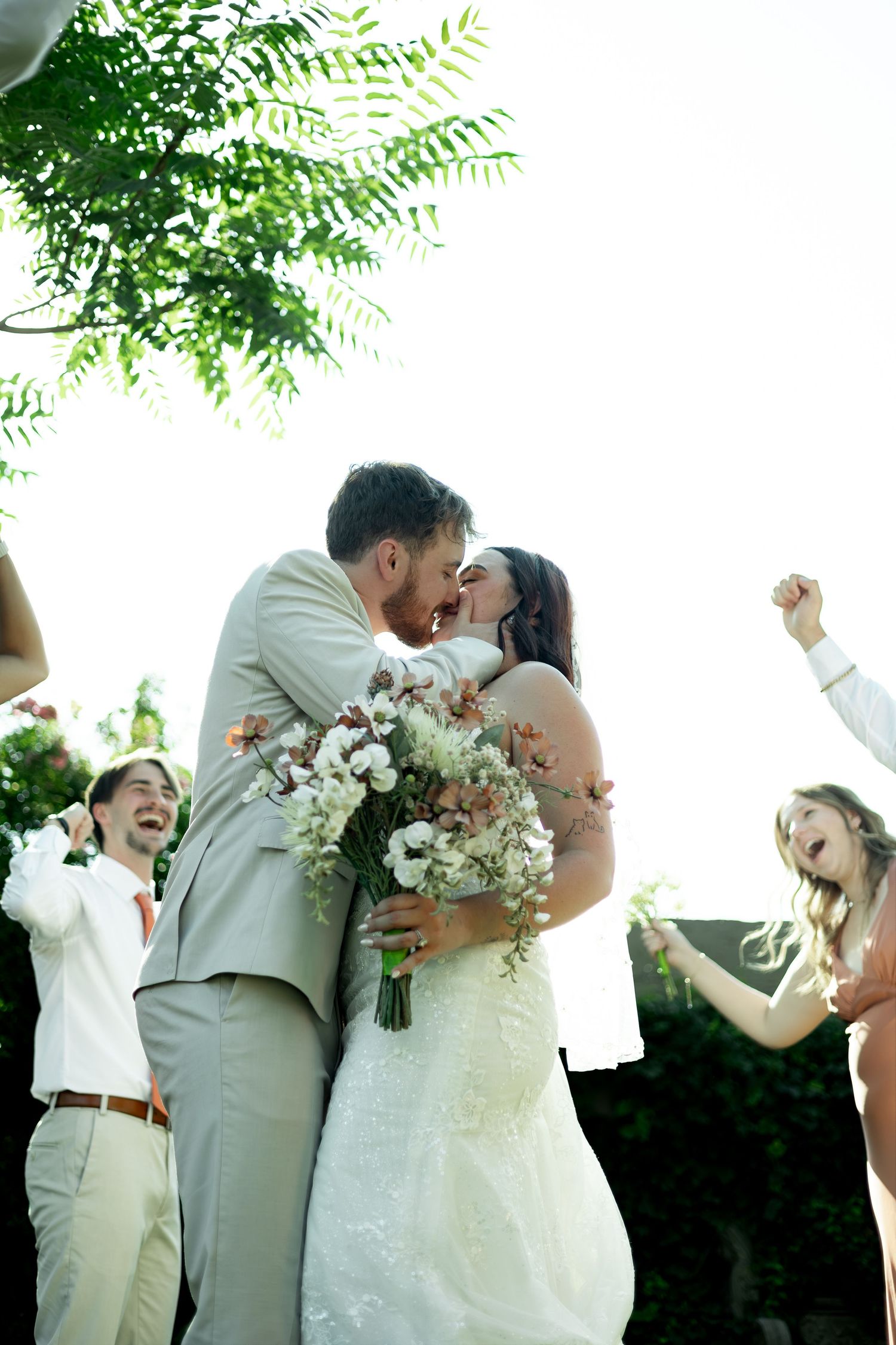 Bride and groom share a kiss while holding a floral bouquet outdoors, as wedding guests cheer around them.
