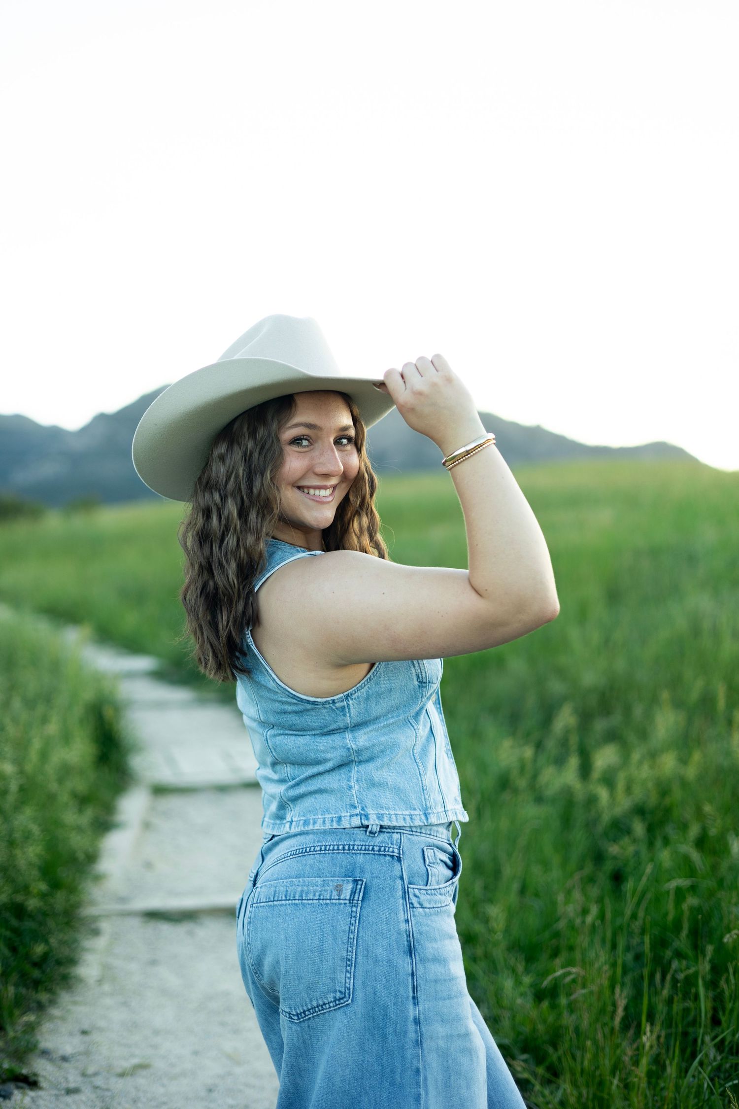 Woman in denim overalls and cowboy hat stands on path through green meadow with mountains in background.