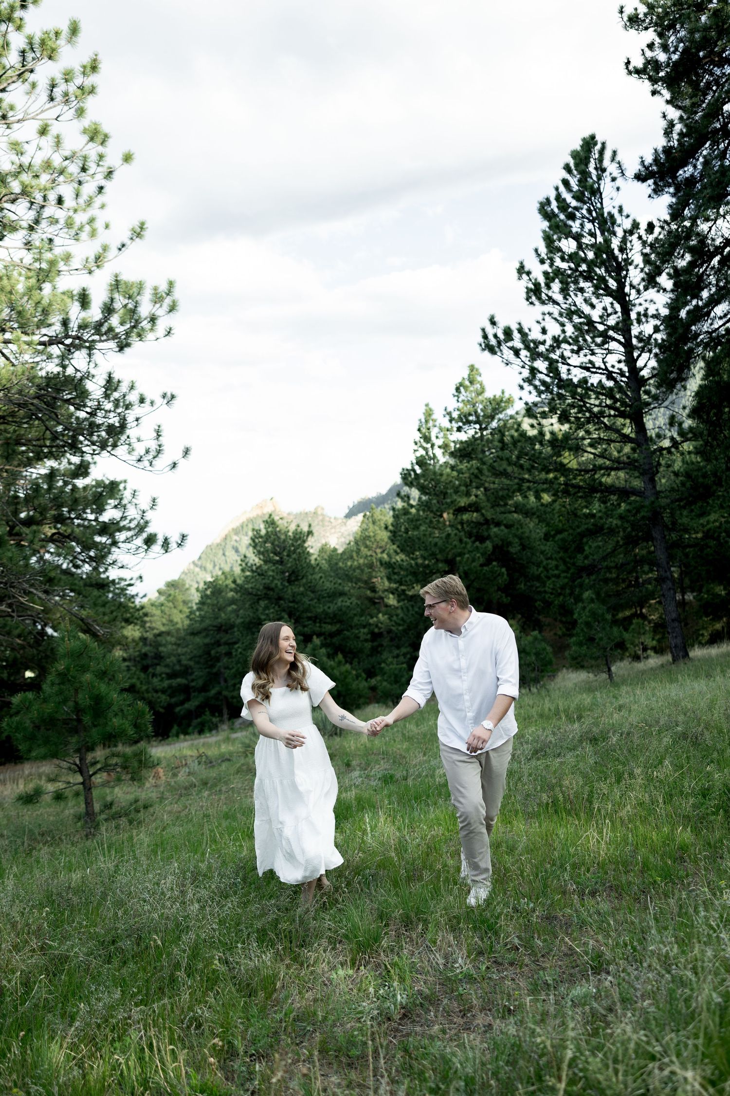 Pregnant woman in white dress holds hands with man in white shirt walking through green mountain meadow with pine trees.