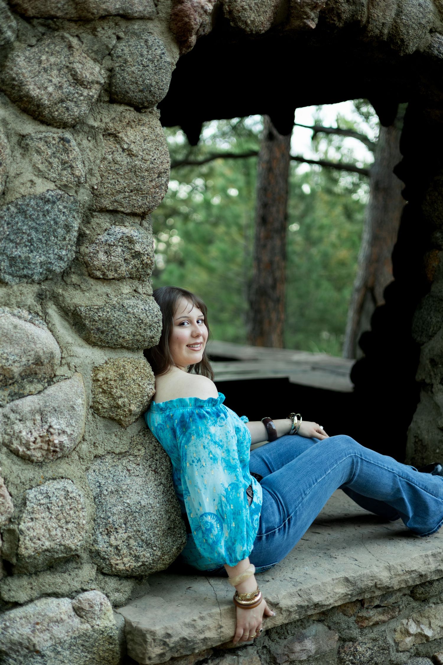 A young woman in a blue off-shoulder top and jeans relaxes against a rustic stone wall outdoors.