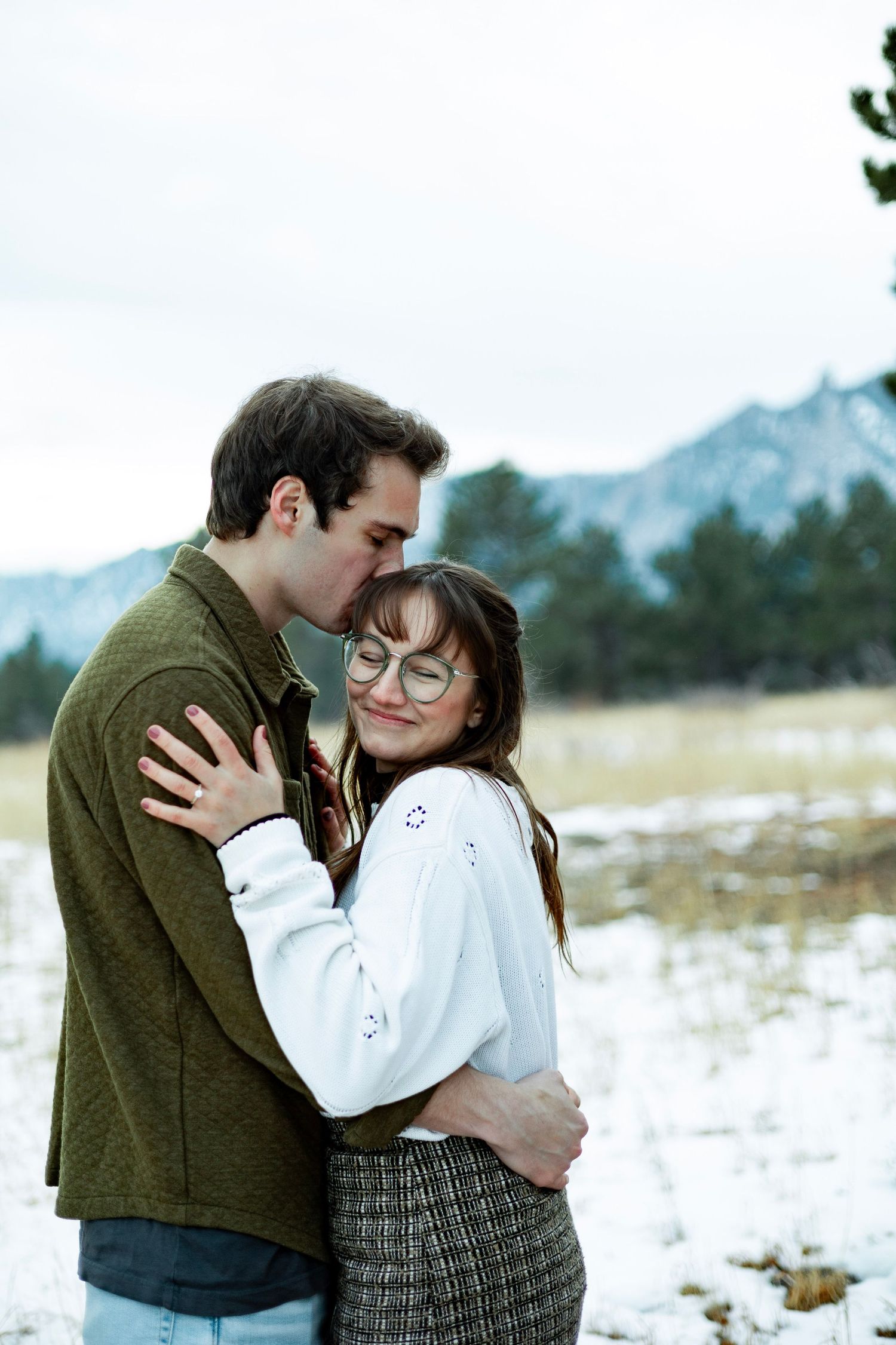 Couple embracing in snowy mountain meadow, man kissing woman's head, she smiles wearing glasses and white sweater.