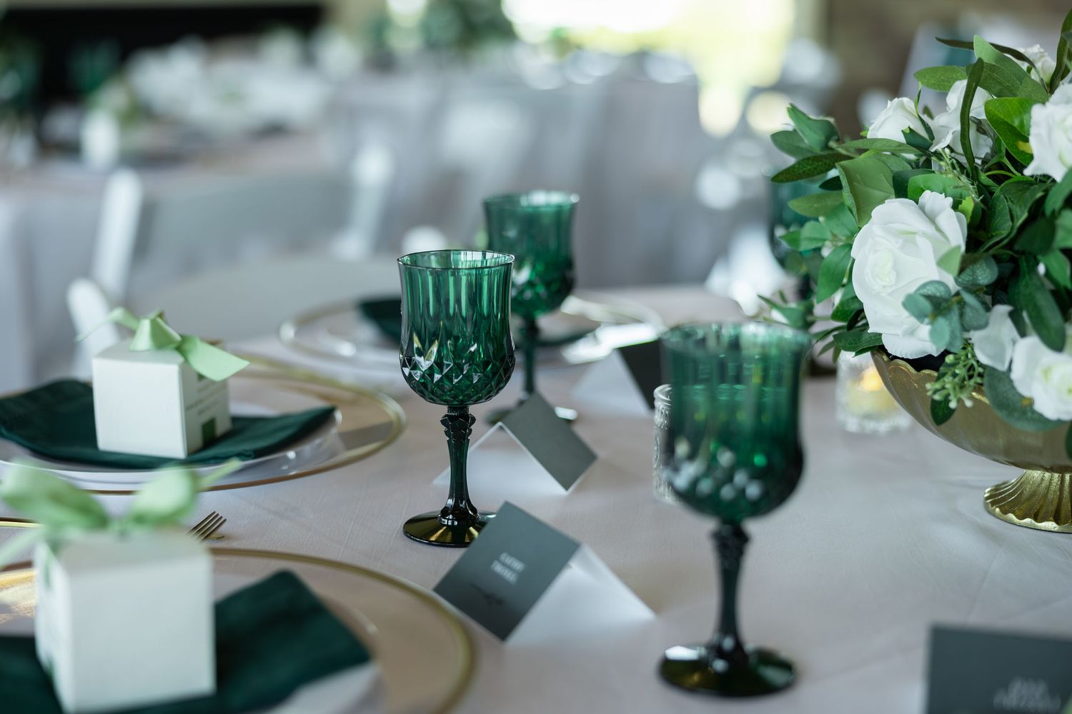 Elegant wedding table setting with emerald green goblets, gold chargers, white floral centerpiece, and dark green linen napkins.