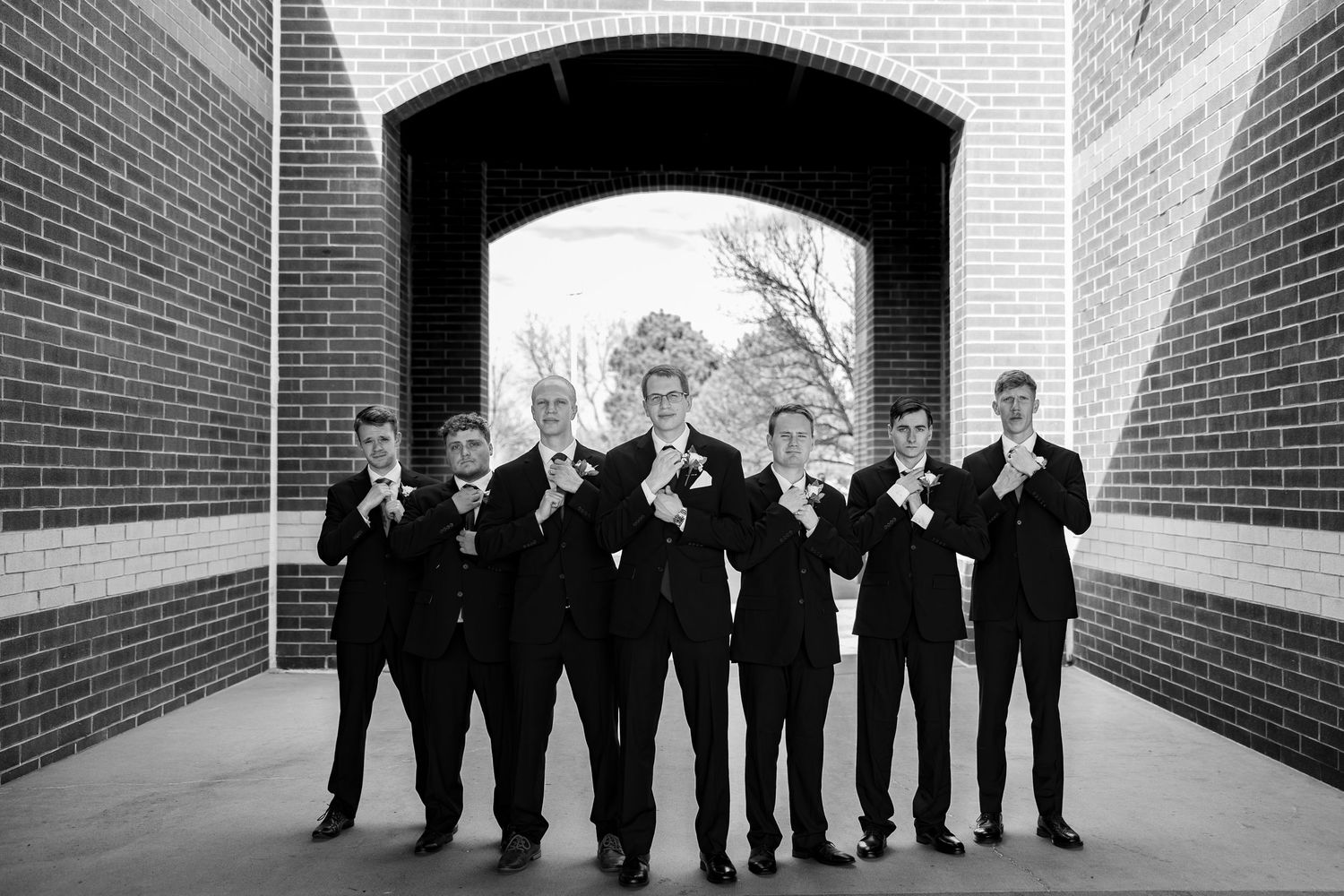 Black and white photo of groomsmen in suits adjusting boutonnieres under a brick archway.