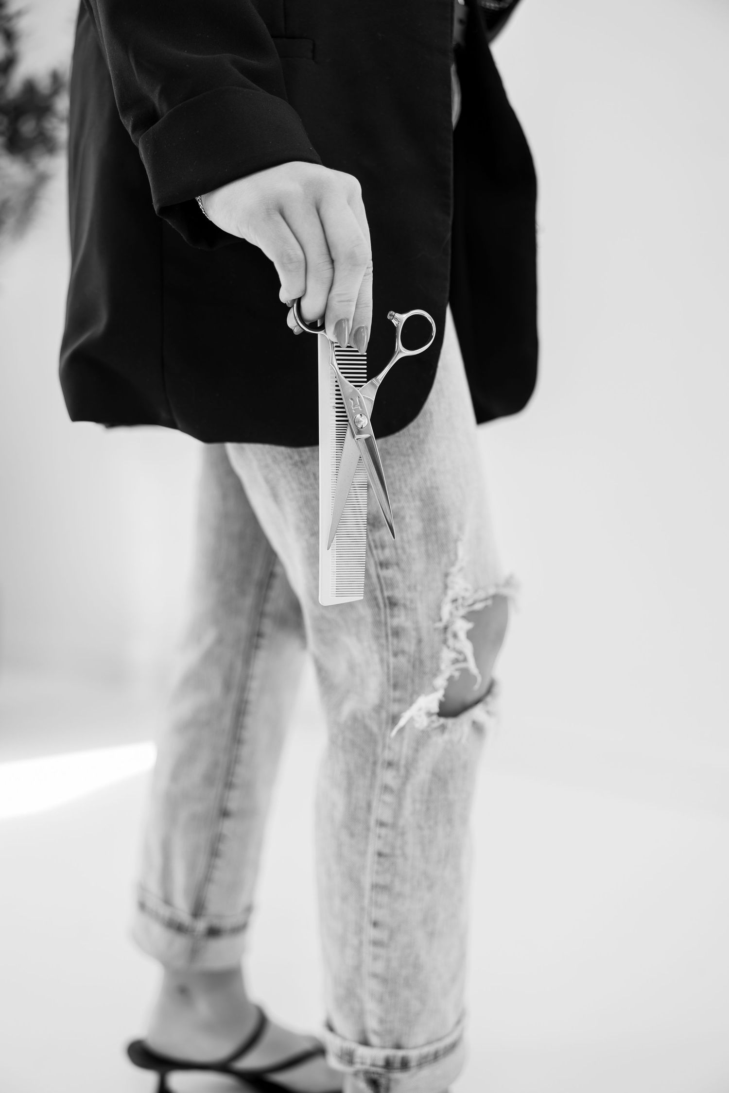 Black and white photo of a hairstylist holding scissors and a comb, wearing ripped jeans and a black blazer.